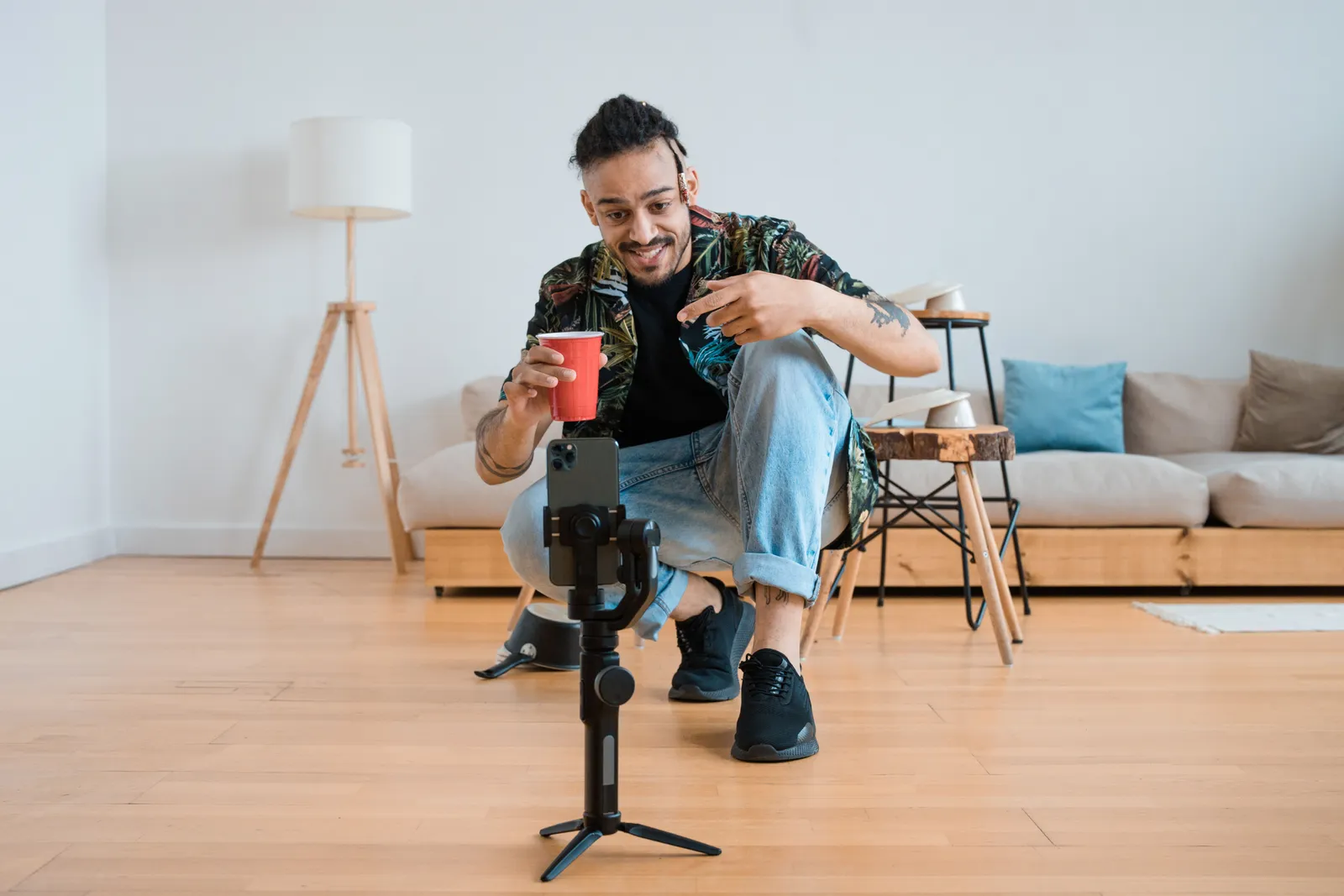 Man filming vlogs in a living room setting, holding a red cup and gesturing at smartphone on tripod