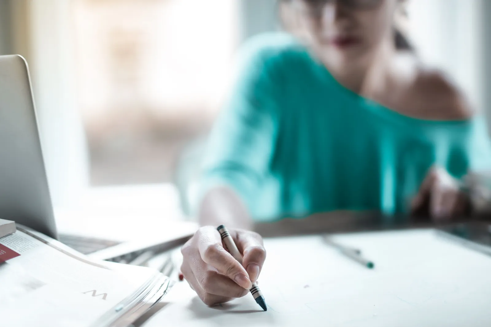 Woman concentrating on sketching at a desk with a laptop and papers in an indoor office setting