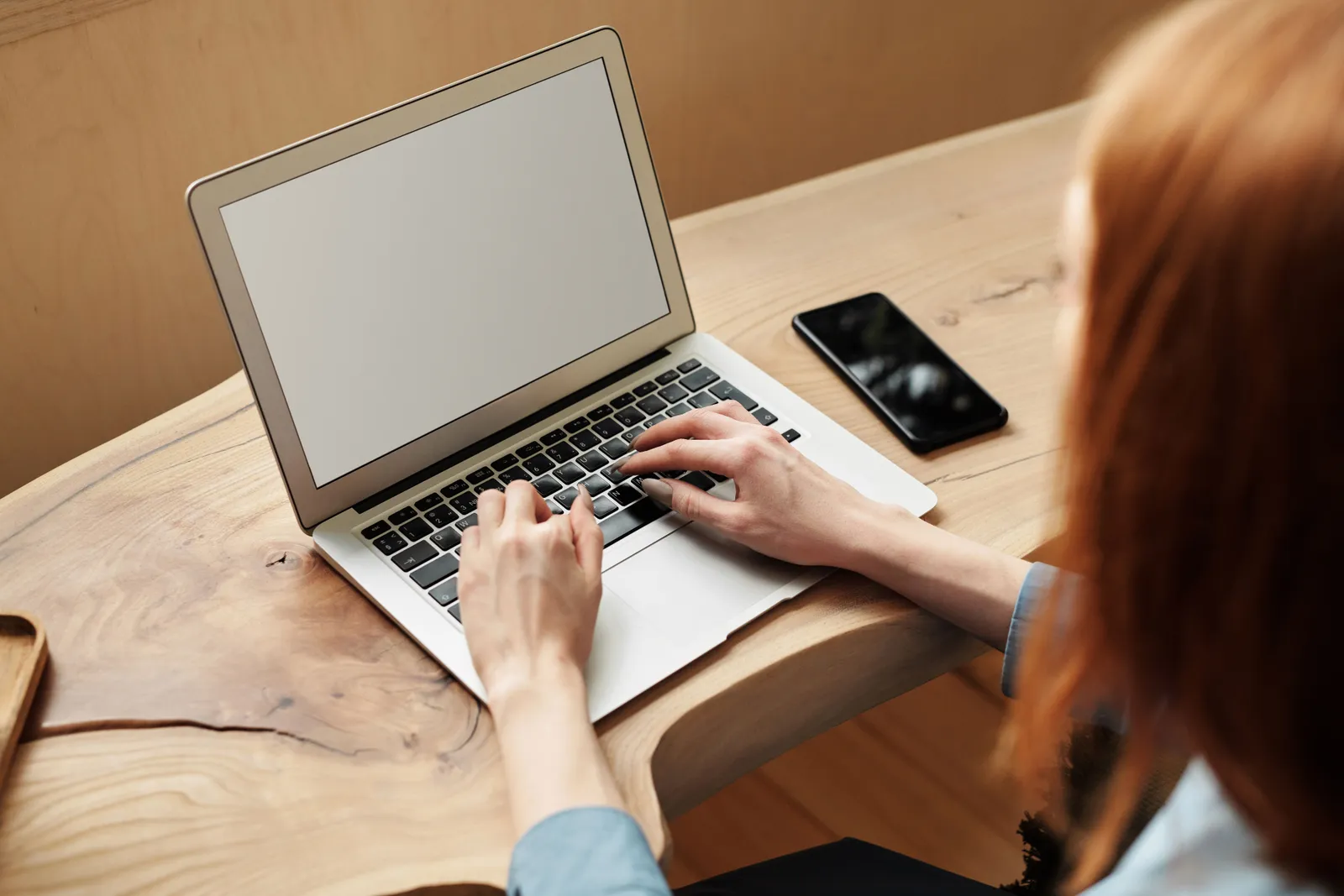 A woman working on a laptop at a wooden desk, indoors with natural light