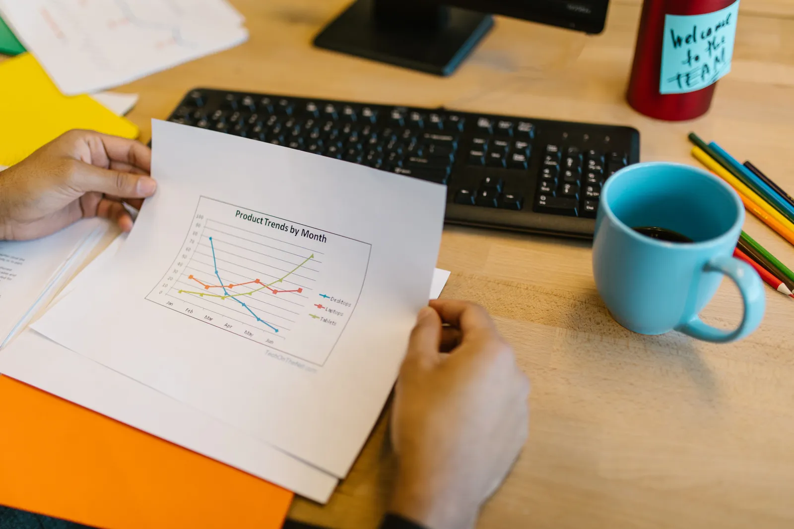 Close-up of hands reviewing a paper graph with a coffee cup and keyboard on a desk