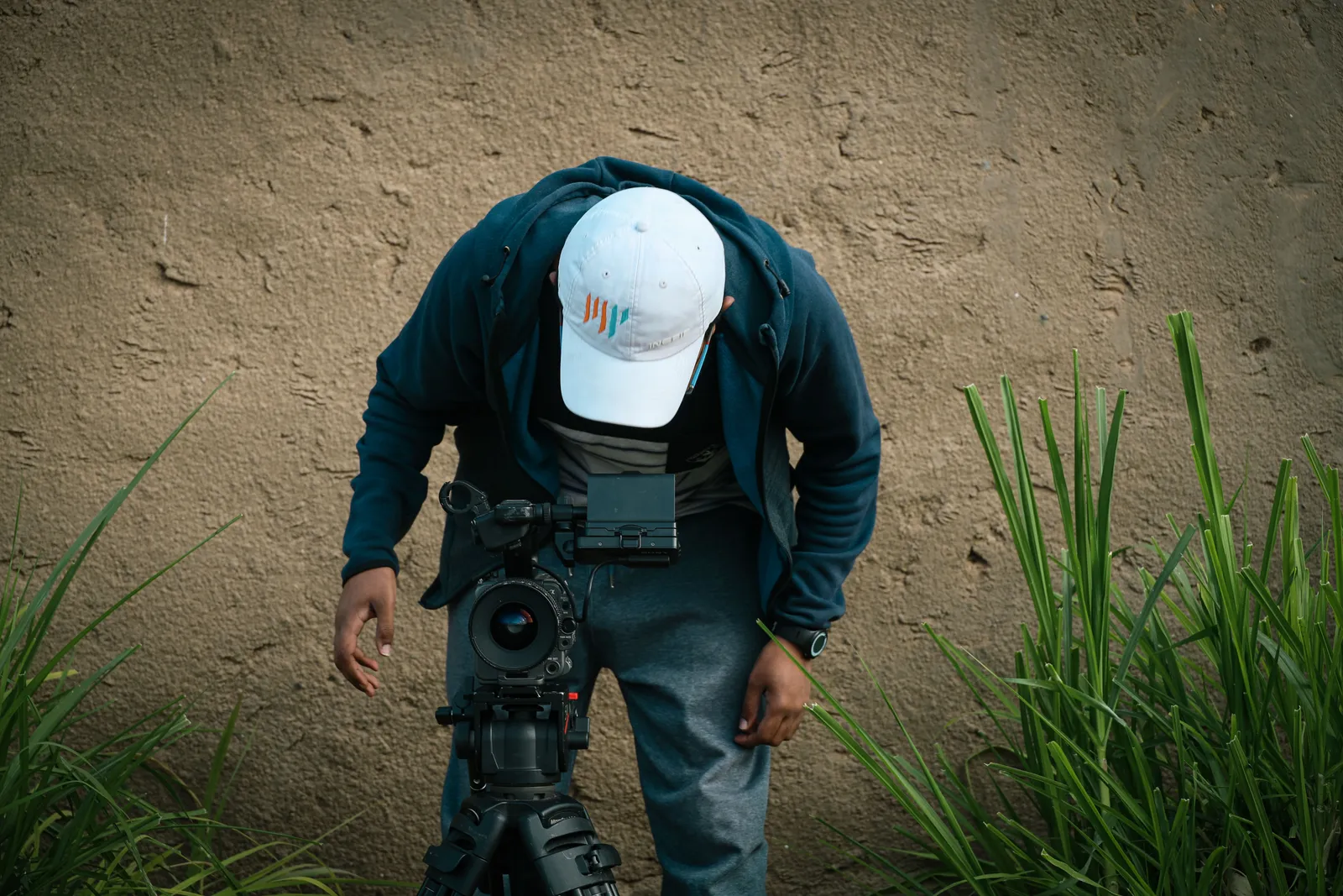A man adjusts a camera setup outdoors near a grassy wall
