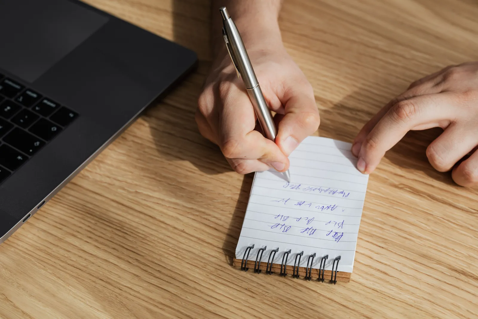 From above of crop unrecognizable man writing out information in spiral notebook while working on business plan on laptop