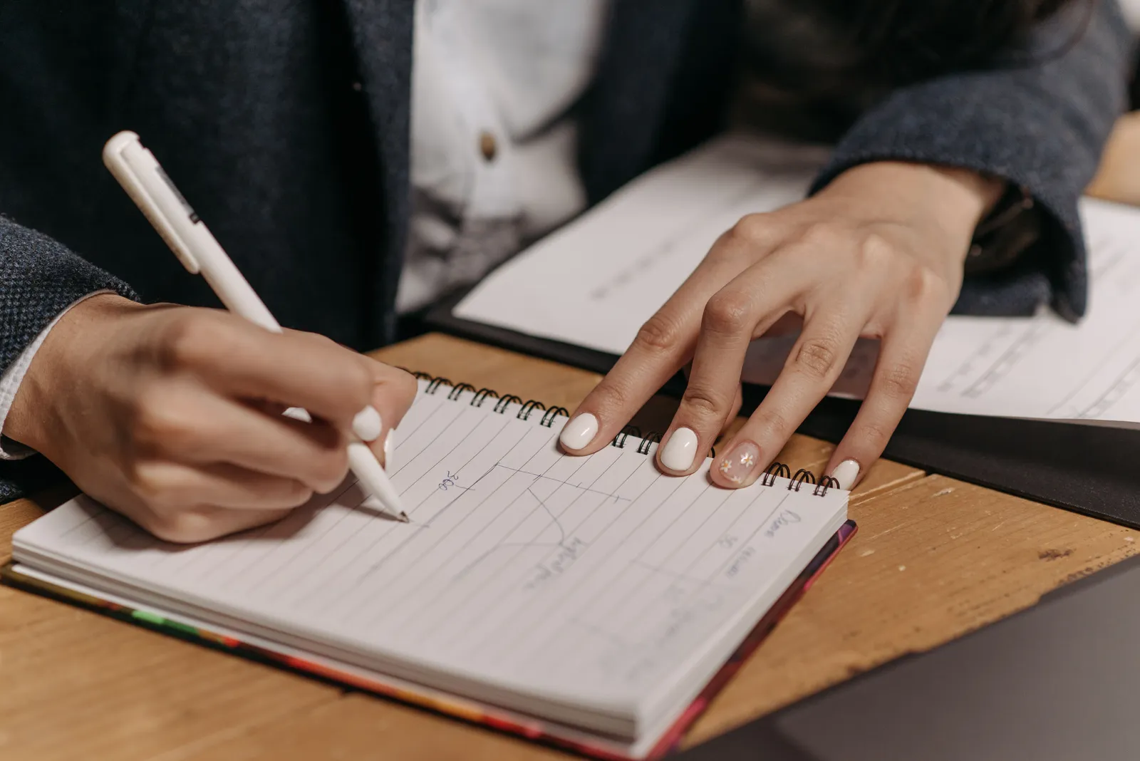 Close-up of a person writing notes in a spiral notebook with a pen on a wooden desk