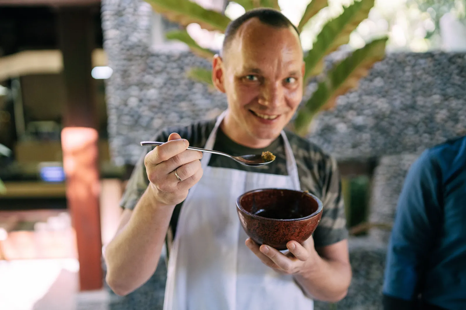 A cheerful man in an apron tasting food from a bowl with a spoon indoors