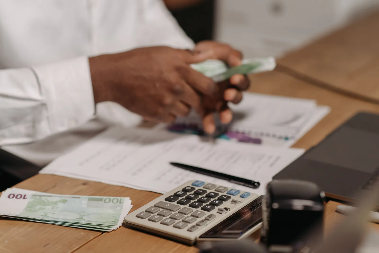 Hands counting euro bills on a wooden desk with calculator, financial documents, and laptop nearby