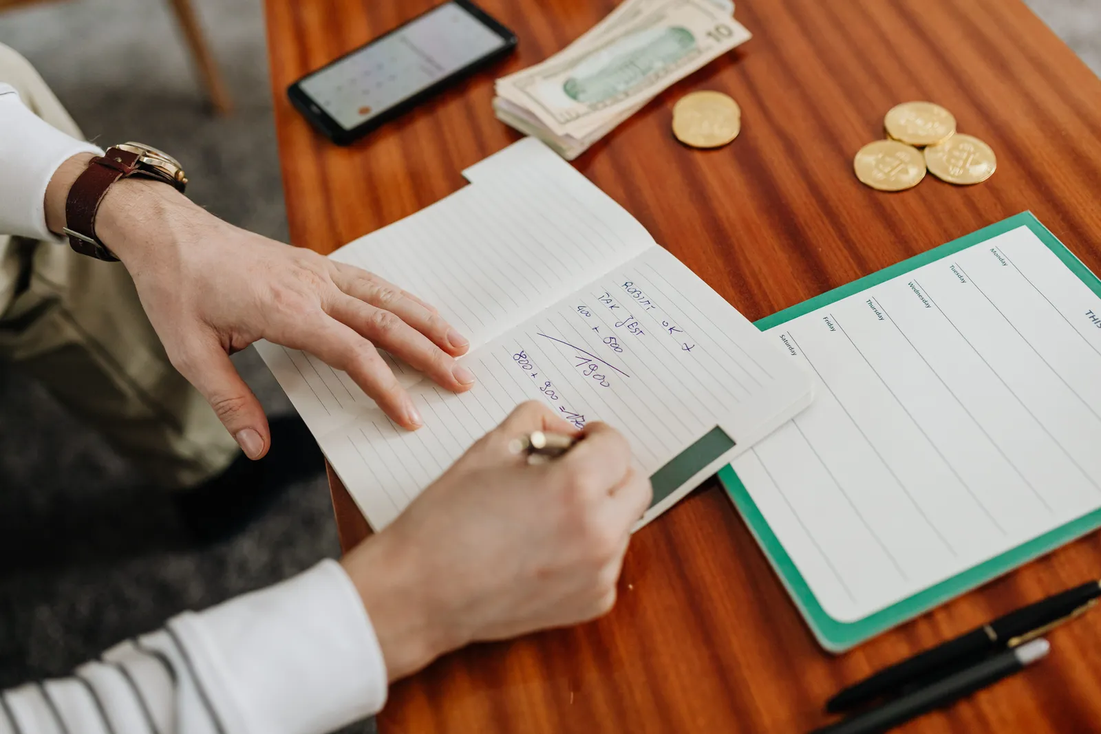 Hands writing financial calculations on notebook with money and coins on table