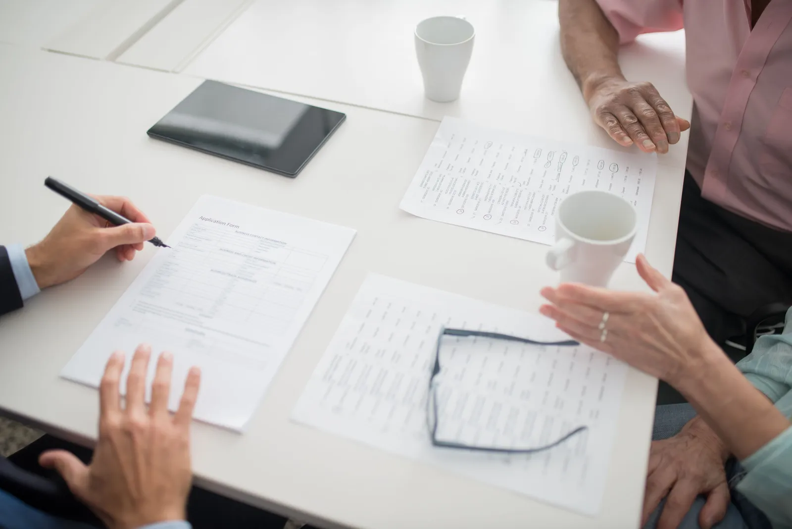 Hands writing on documents in a business meeting setup with coffee cups and tablet