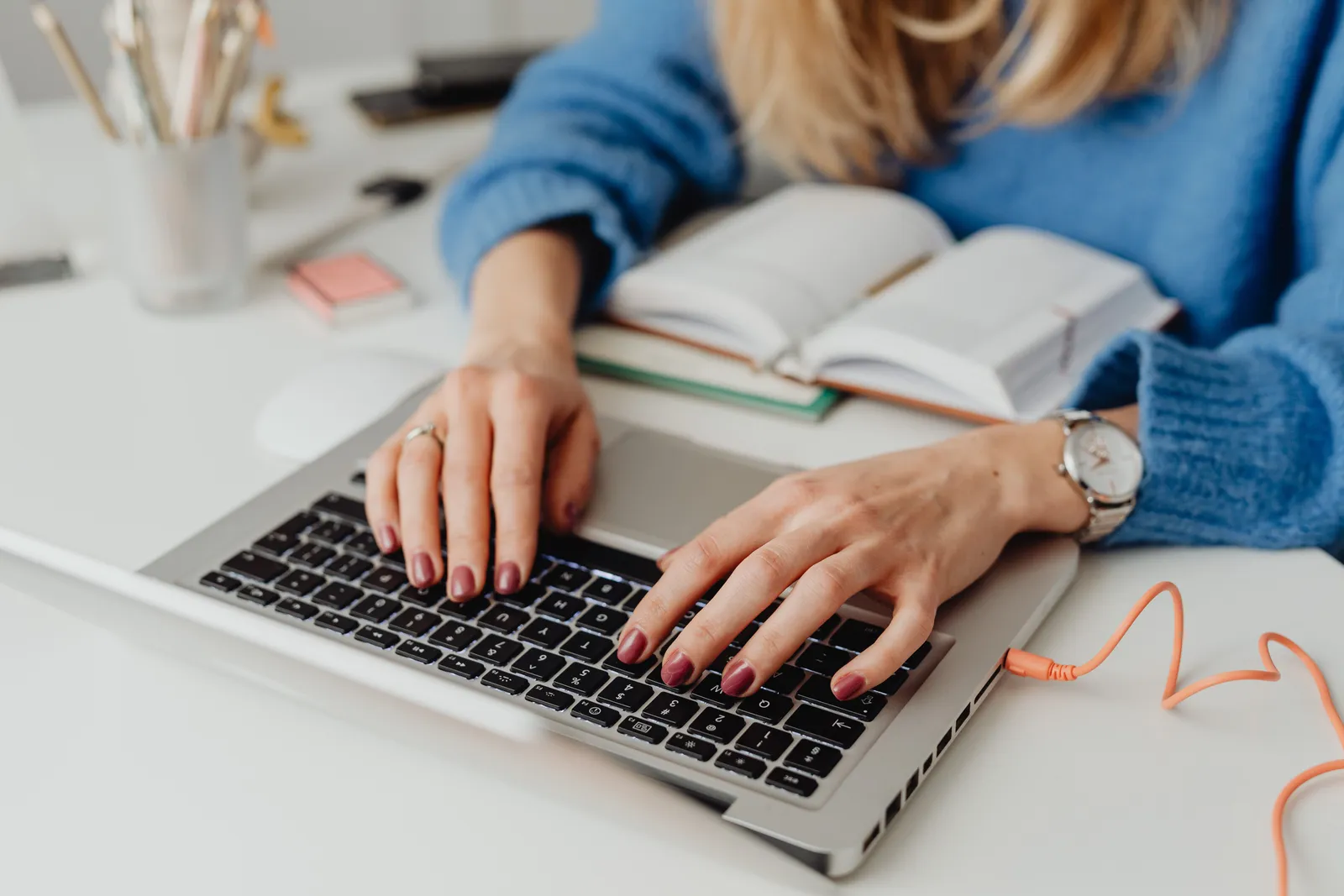 Close-up of hands on laptop keyboard, alongside an open book and stationery tools, in an indoor setting
