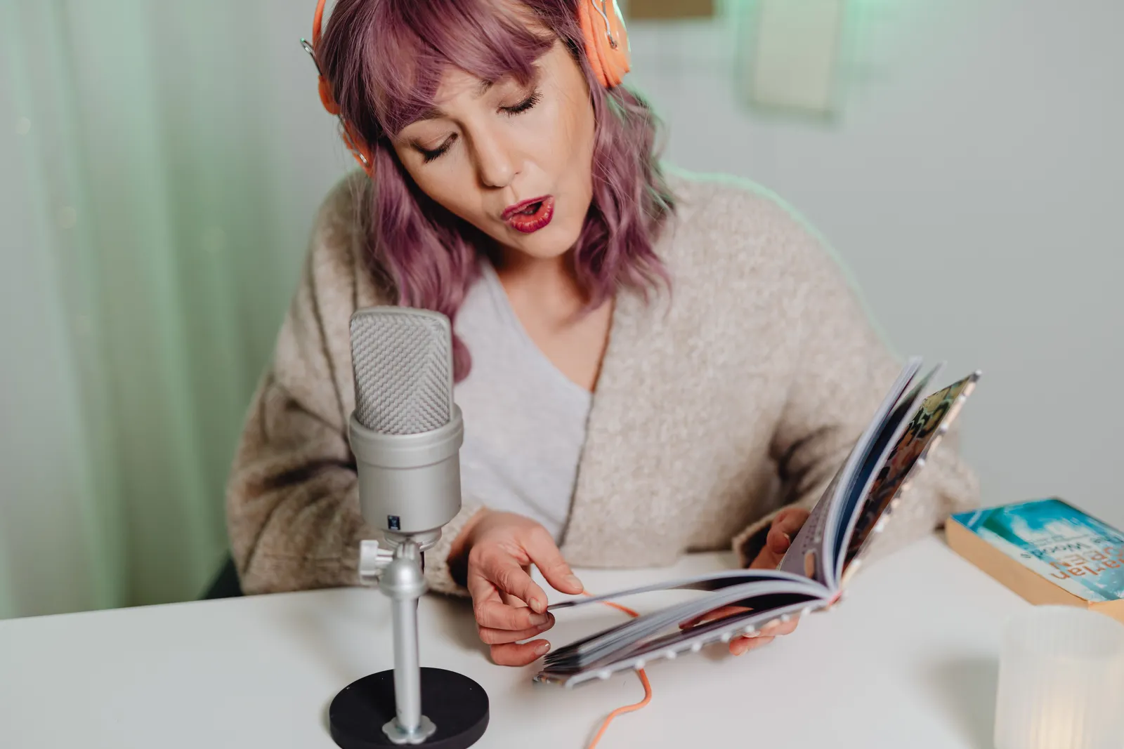 Woman with pink hair singing while reading a book, wearing headphones, seated at a white table with a microphone