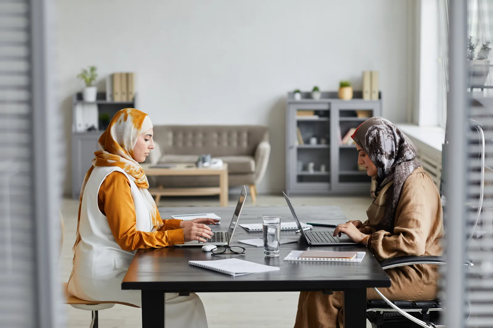 Two women wearing hijabs working on laptops at a modern desk in an office setting