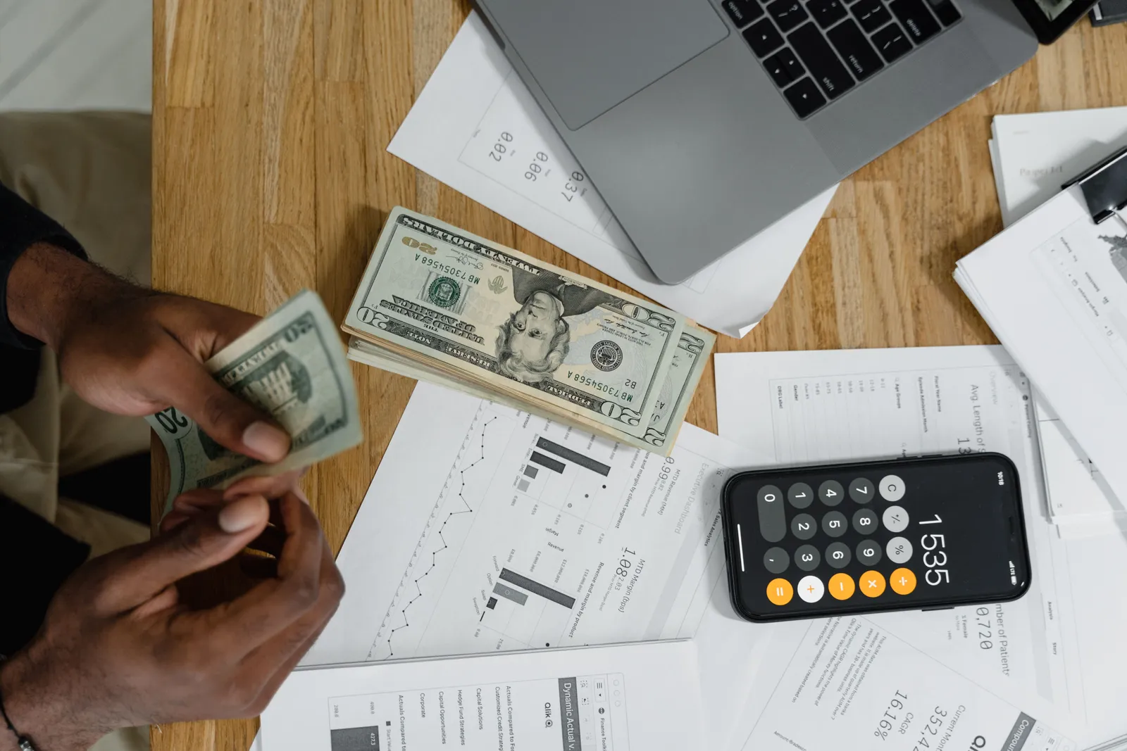 Close-up of hands counting cash on desk with calculator, charts, and laptop, illustrating financial management
