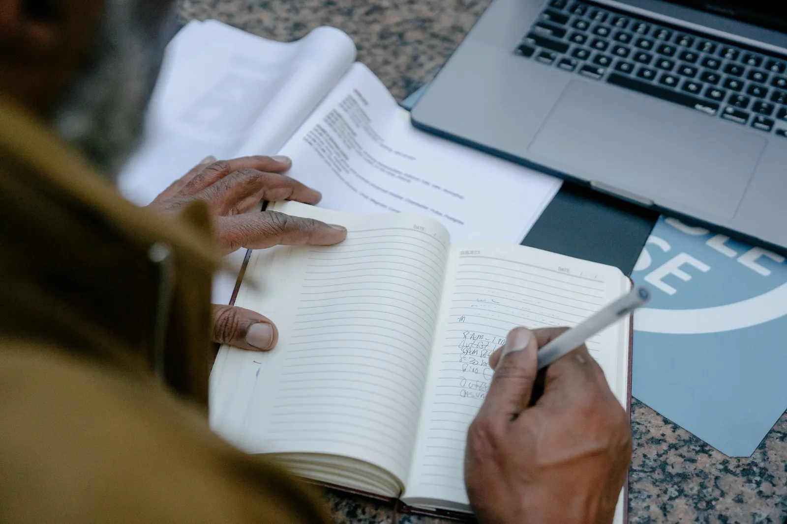Businessman writing in notebook at desk with laptop and documents