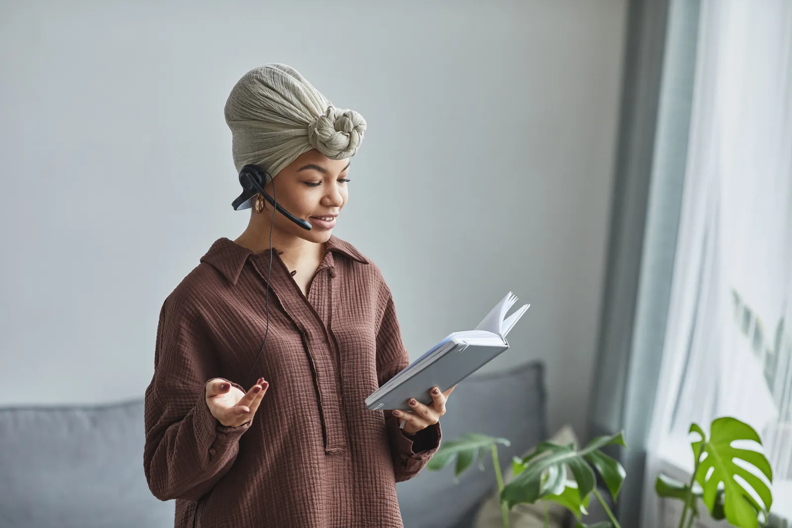 Positive female call agent with microphone speaking while reading notes in notepad during work in light room at home
