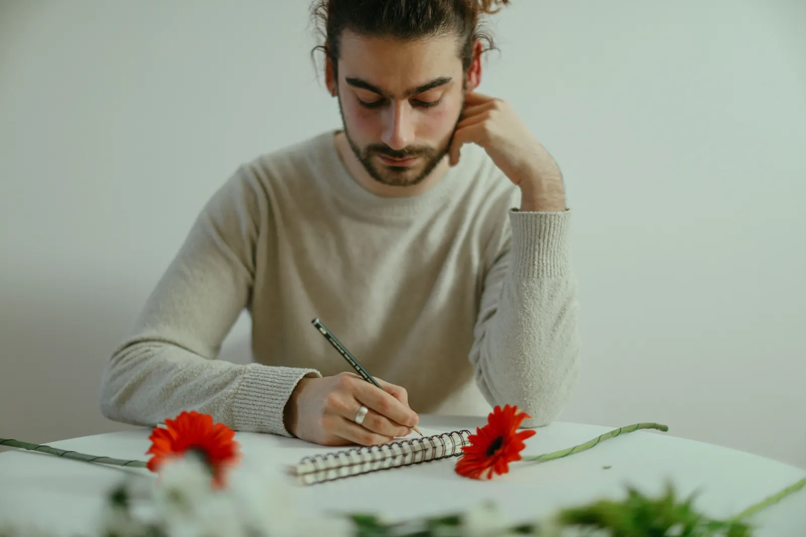 A focused man writing in a notebook, surrounded by red flowers on a table in an indoor setting
