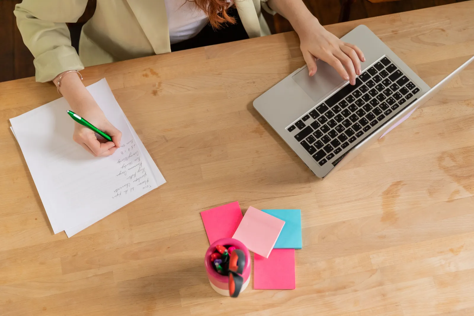 Overhead view of a person writing notes and typing on a laptop at a wooden desk