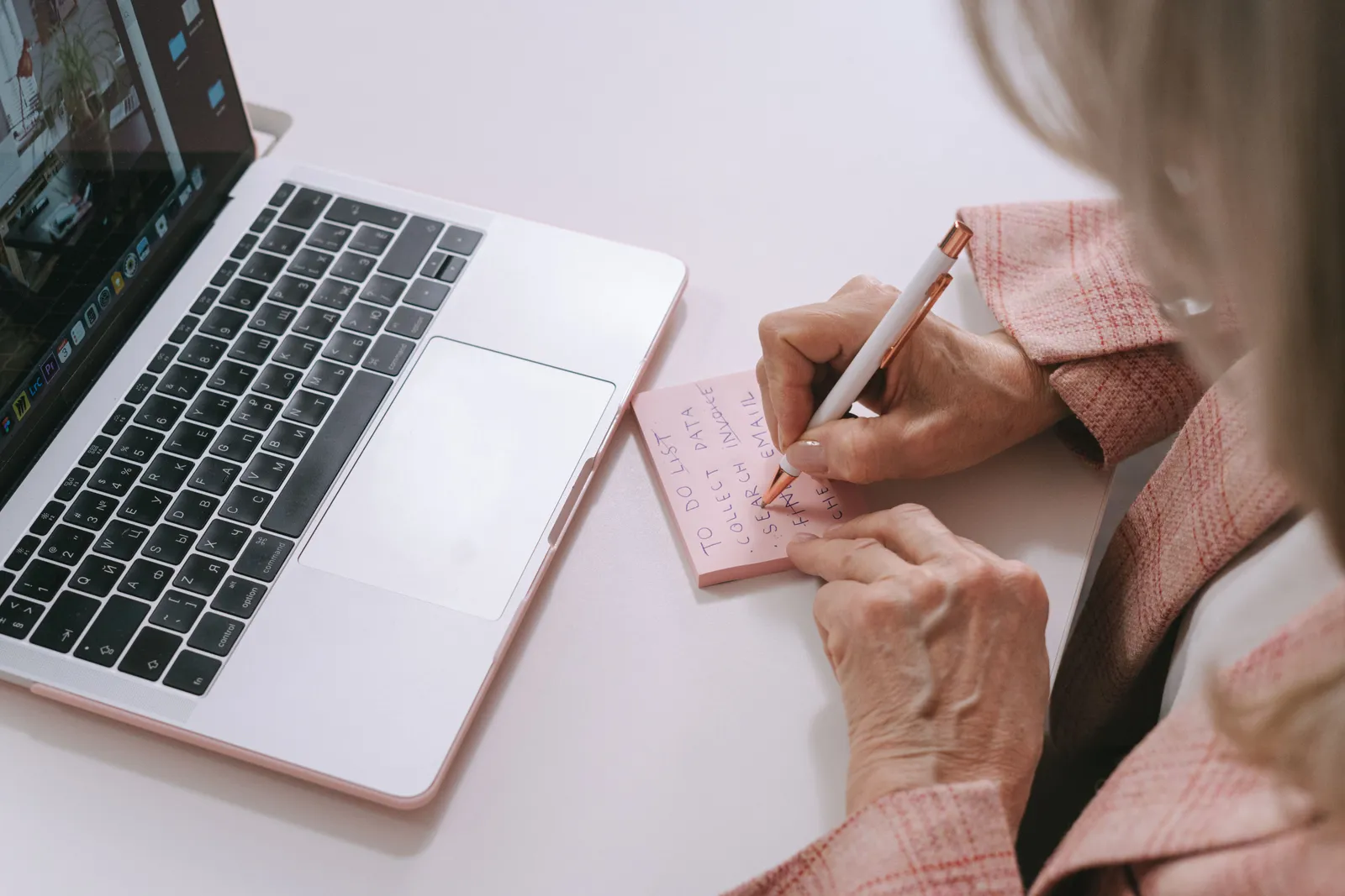 Close-up of a woman writing on a pink sticky note beside a laptop. Productive work setting