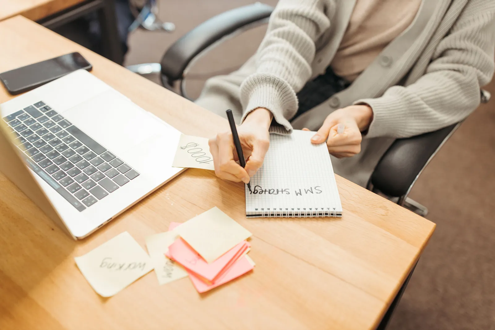 Adult writing notes in a notebook at a desk with a laptop, emphasizing productivity