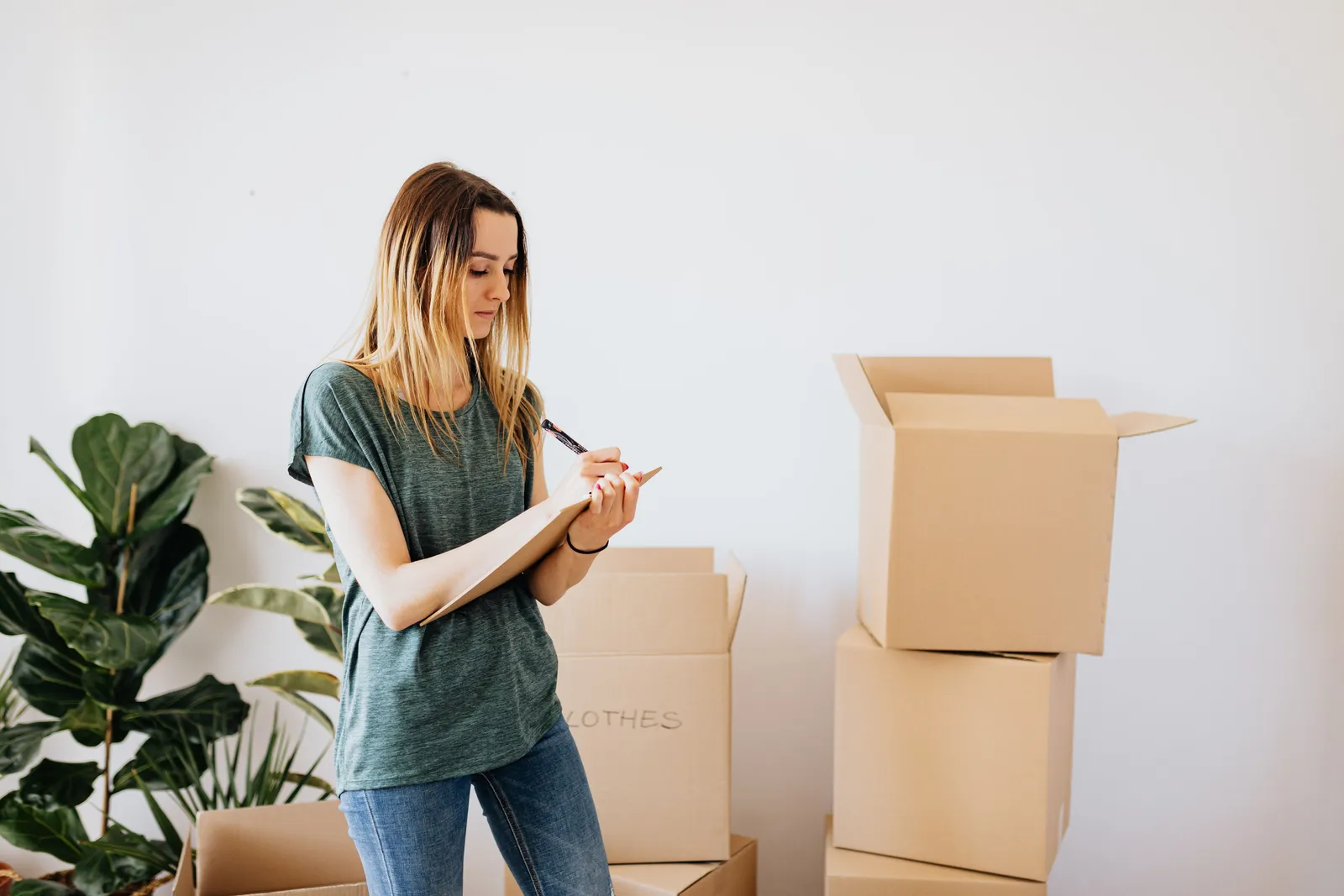 Focused young lady in casual wear taking notes in clipboard while standing near packed carton boxes before moving into new