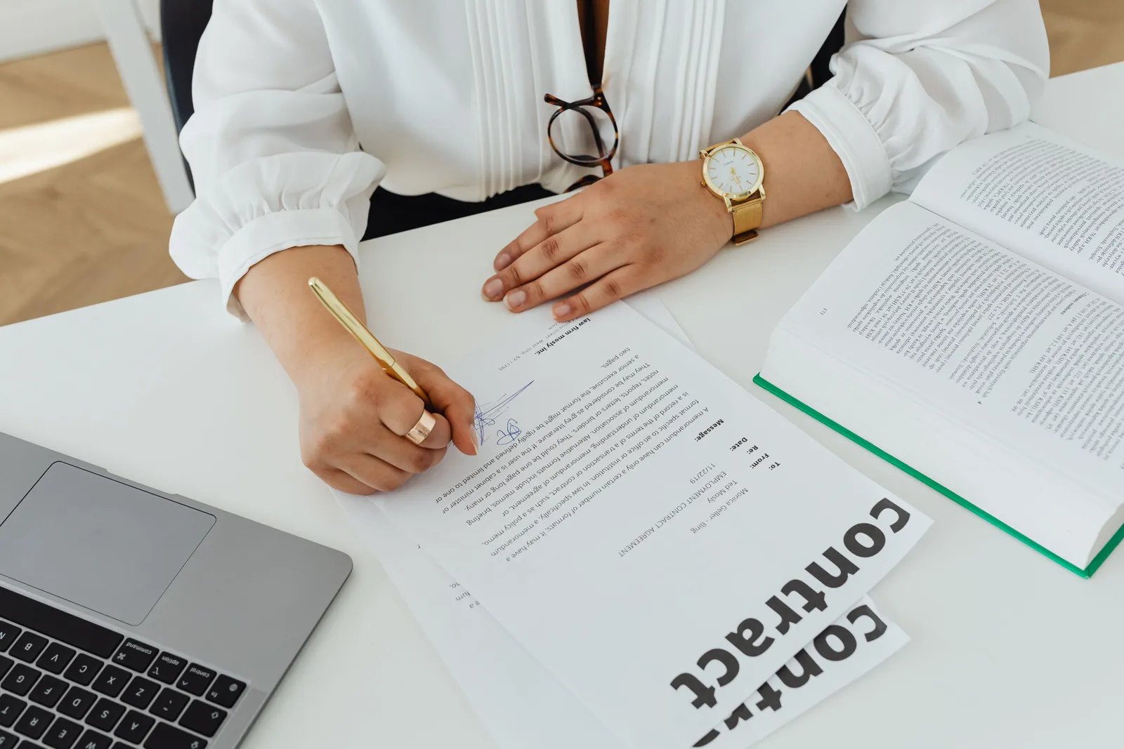 Close-up of a businesswoman signing a contract at her desk, with a laptop and book