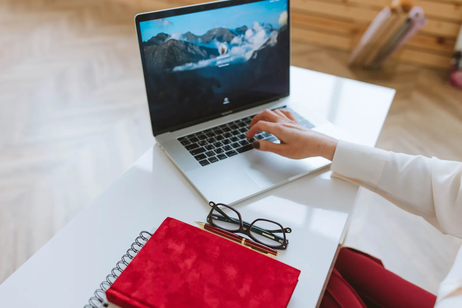 A person working on a laptop with a red notebook and glasses on a white table