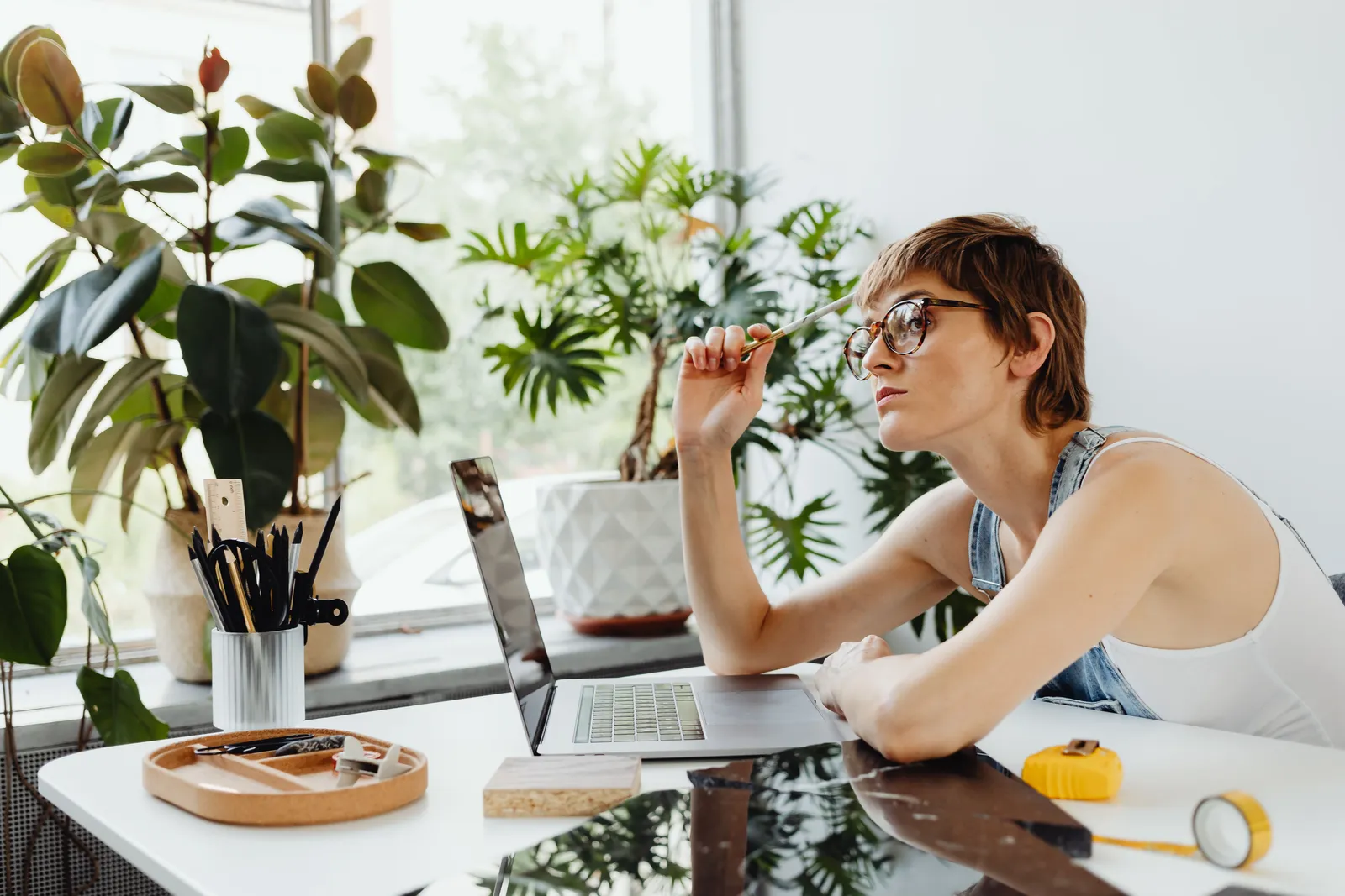 Woman in casual wear working on laptop at desk surrounded by plants. Creative and thoughtful workspace