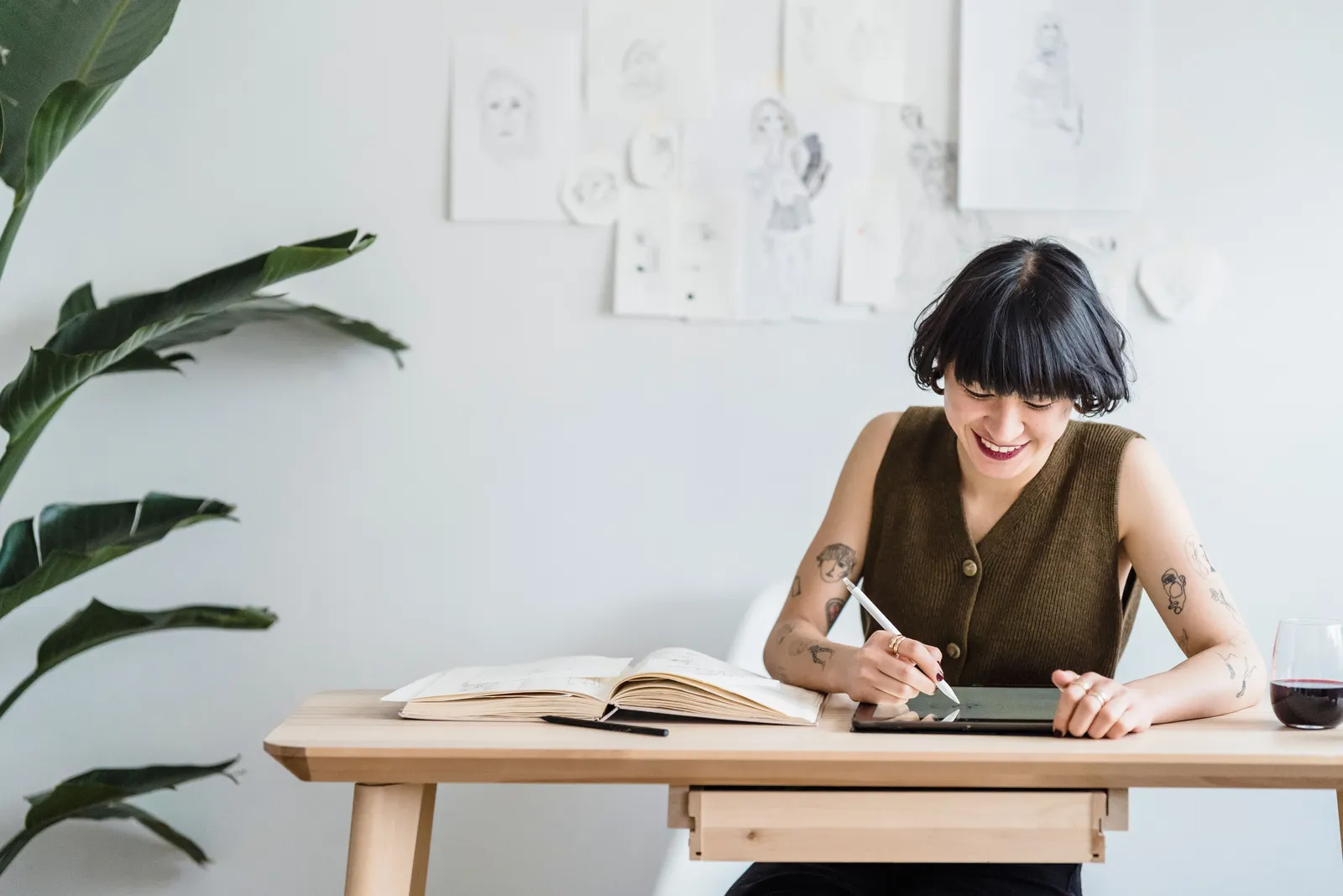 Smiling woman drawing on digital tablet with stylus at a wooden desk, surrounded by sketches