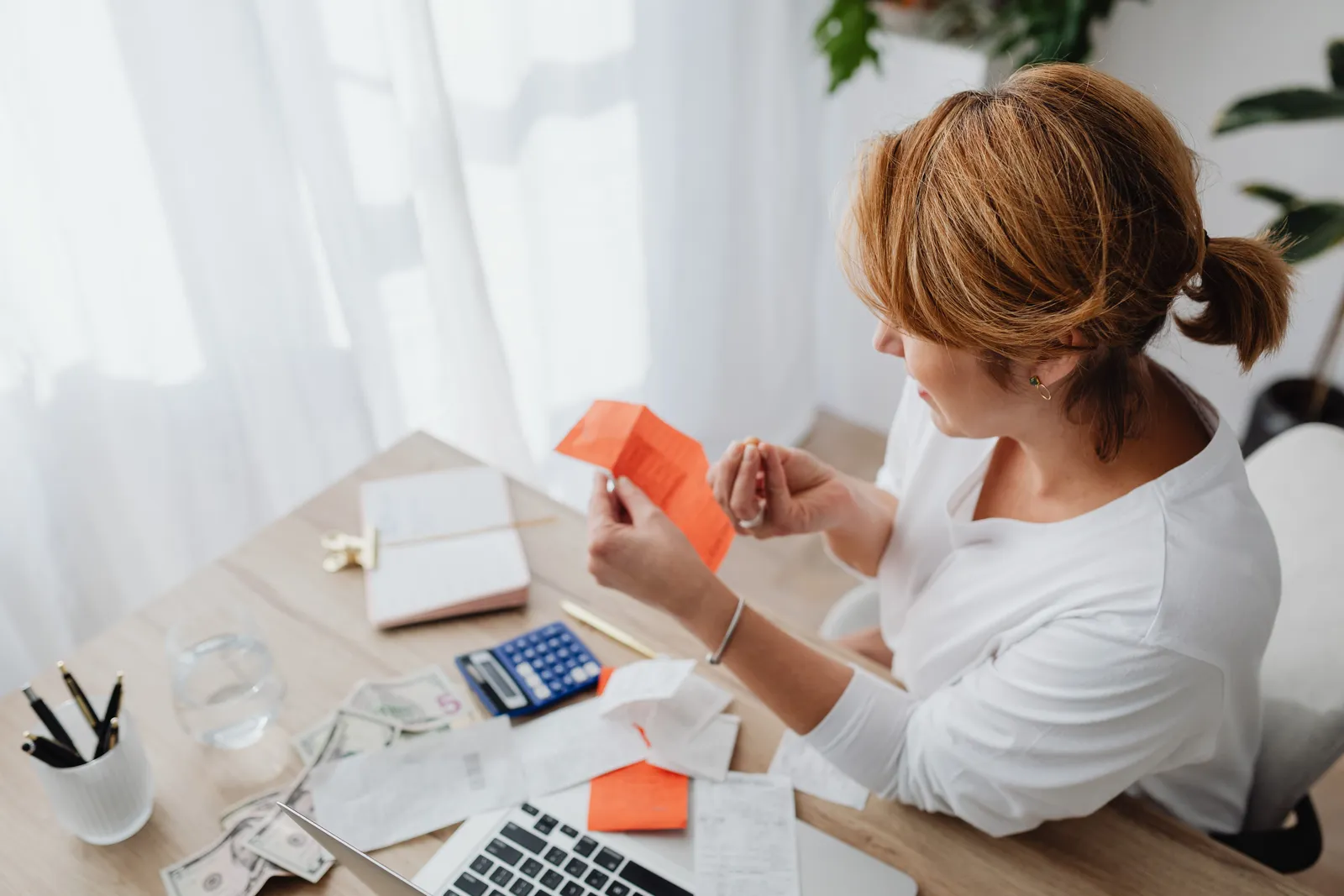A woman organizing her finances with bills and receipts at a home office desk using a calculator and laptop