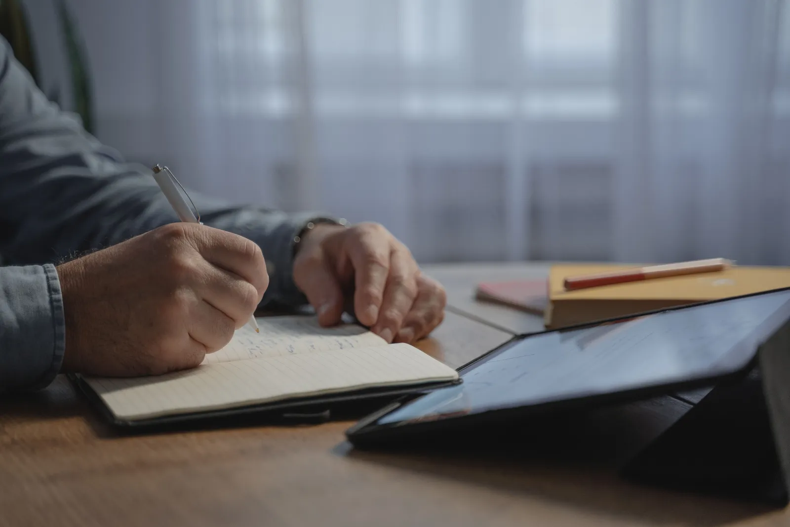 Close-up of a person writing notes in a notebook at a desk with a tablet. Ideal for productivity and workspace concepts