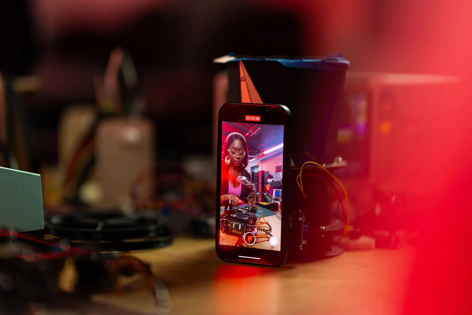A woman records a tech tutorial using her smartphone on a desk filled with electronics