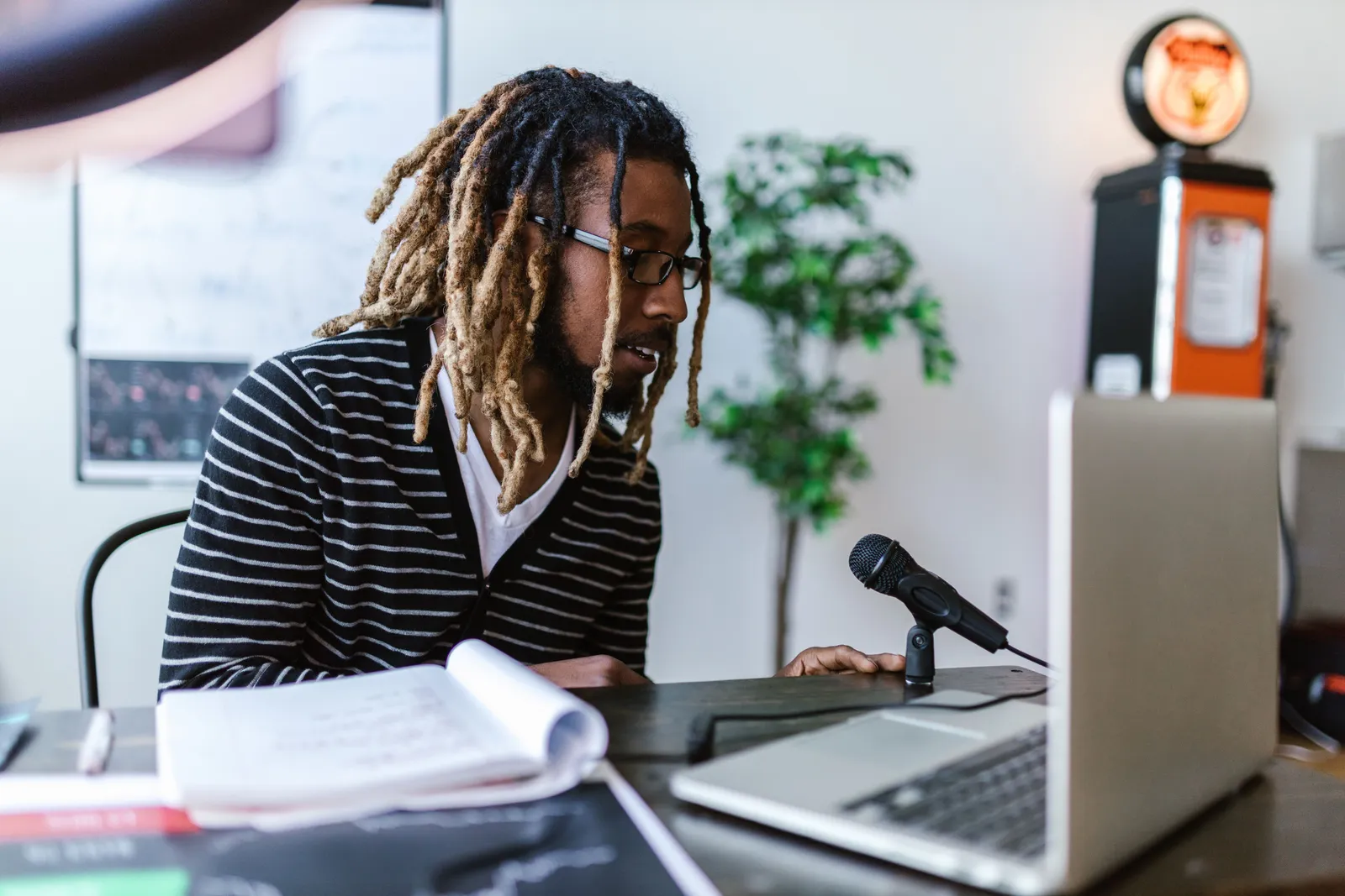 A young man with dreadlocks and eyeglasses records a podcast at a desk with a microphone and laptop