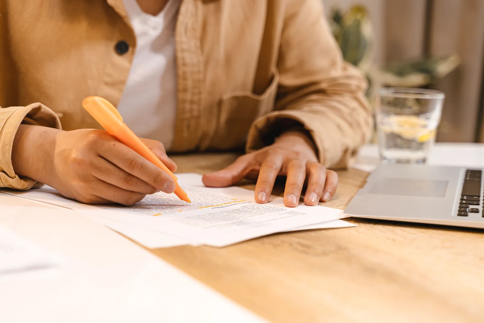 Person highlighting notes at a desk with a laptop and glass of water nearby