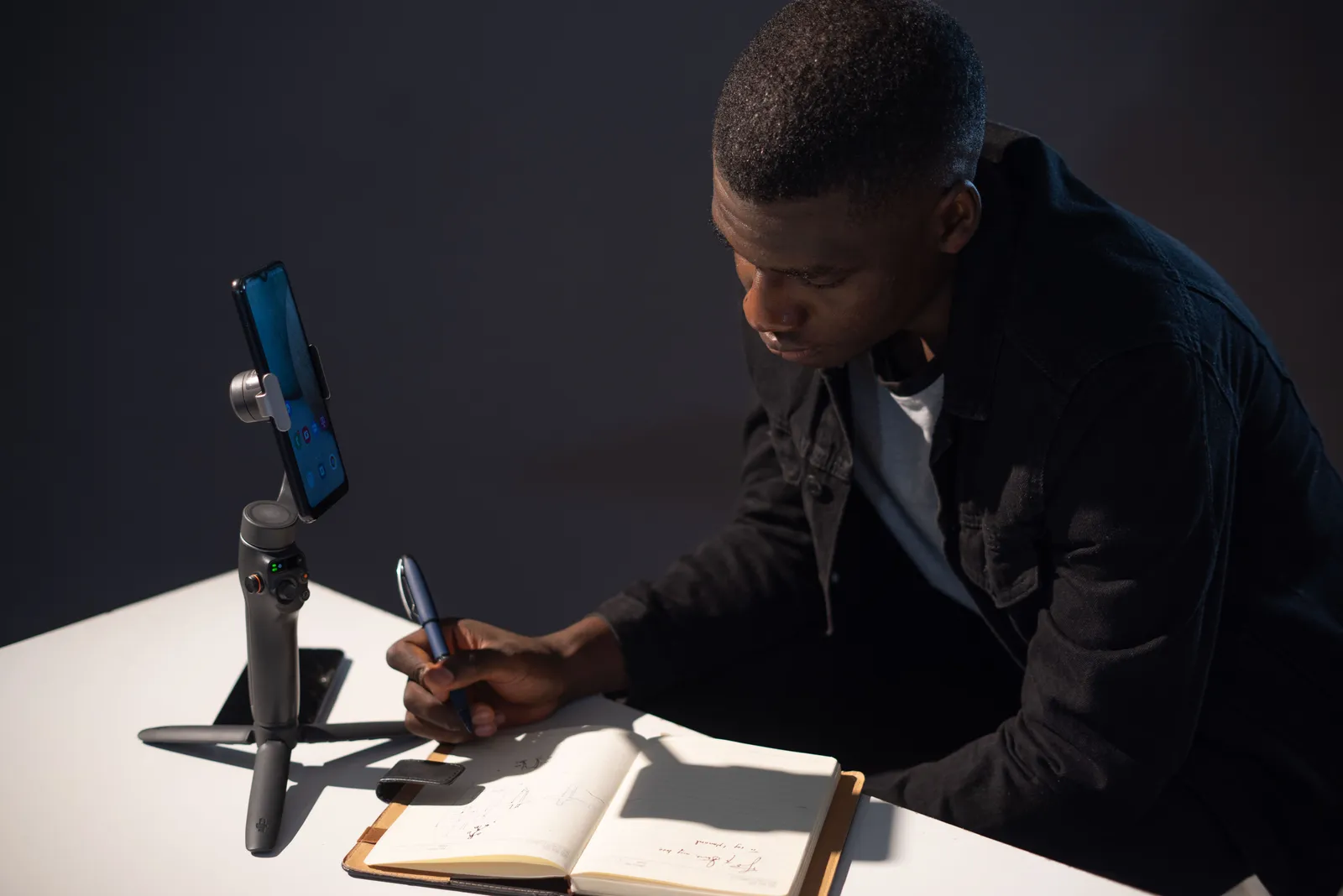 Man writing notes in a notebook with smartphone on gimbal at desk, showcasing remote work