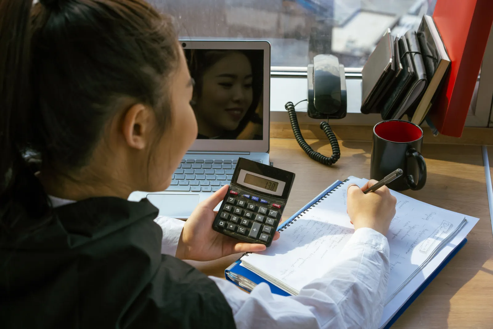 Woman using a calculator at a desk with a laptop and notepad, involved in office work