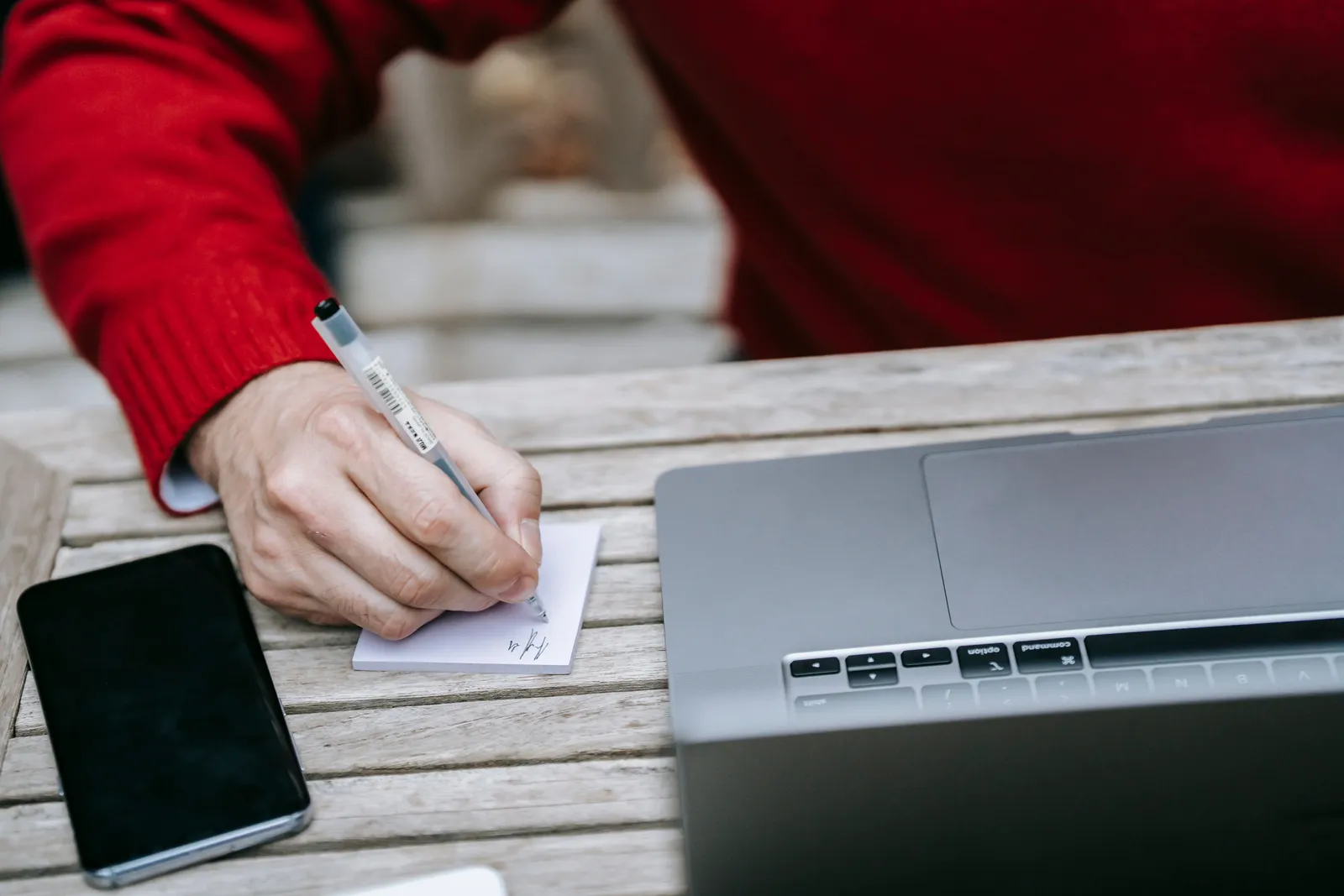 Adult writing notes beside a laptop and smartphone on a wooden table outdoors