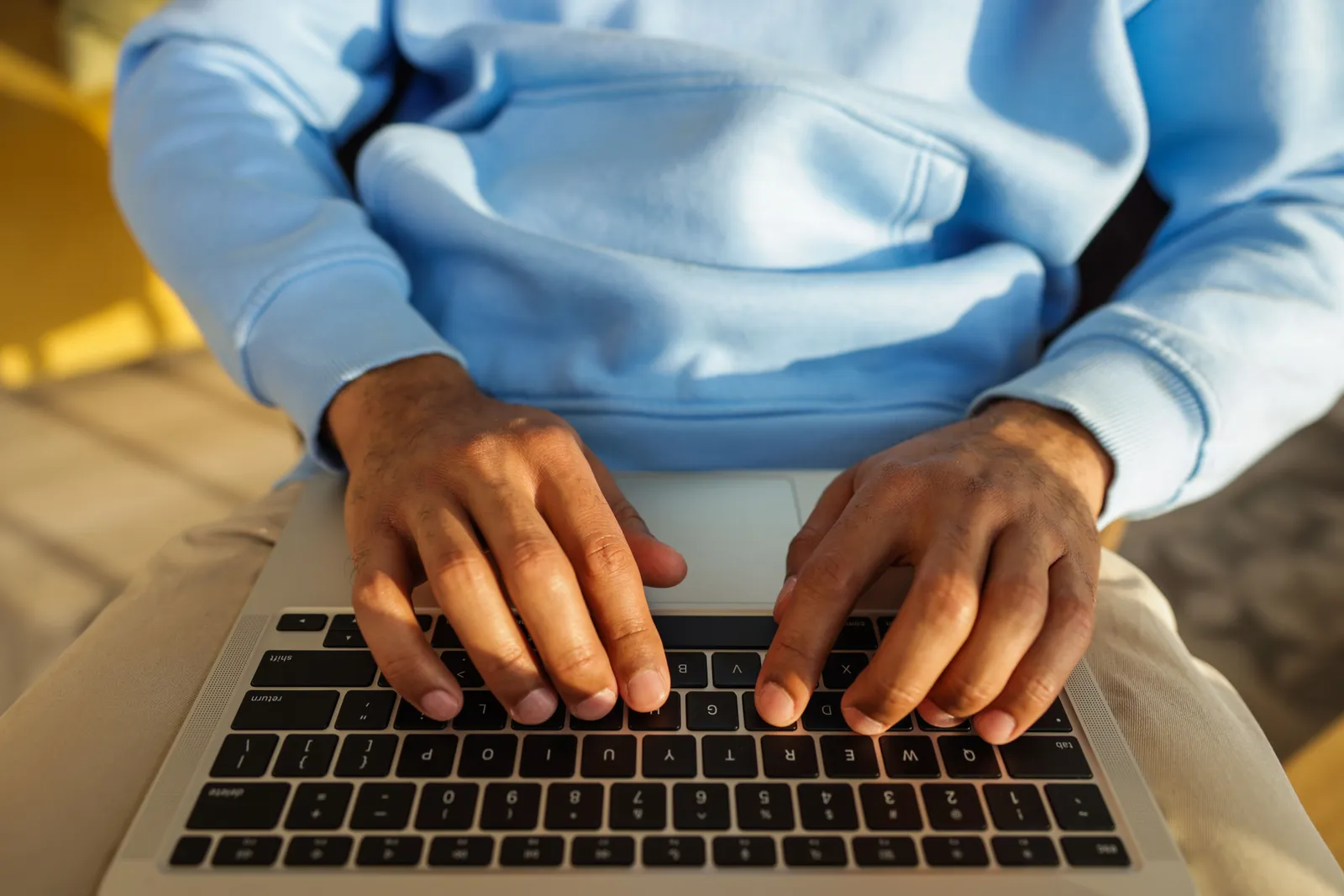 Hands in a blue hoodie typing on a laptop keyboard, indoors, in bright light