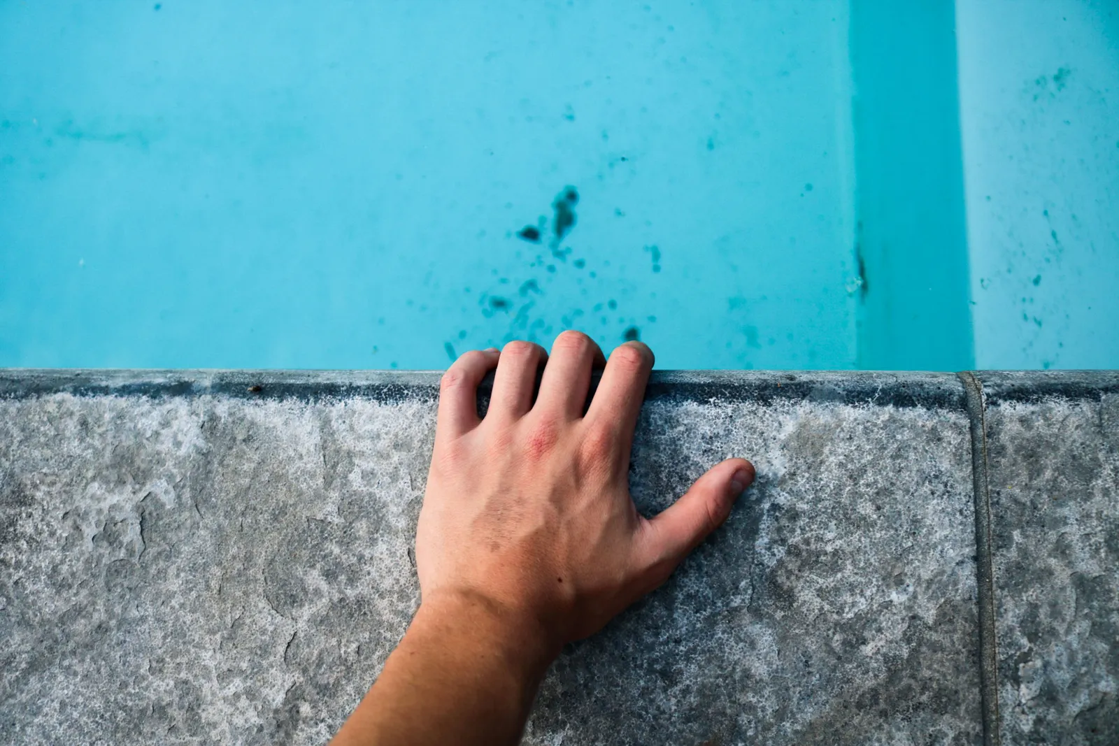 Close-up of a human hand on a concrete edge by a pool with clear blue water