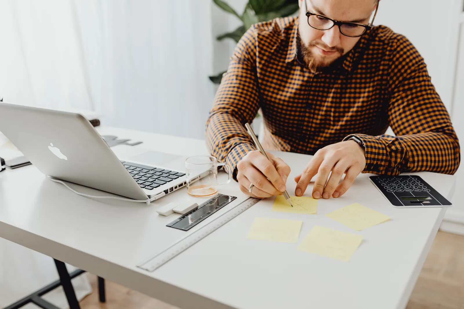 A man with glasses writes notes at his office desk with a laptop and coffee