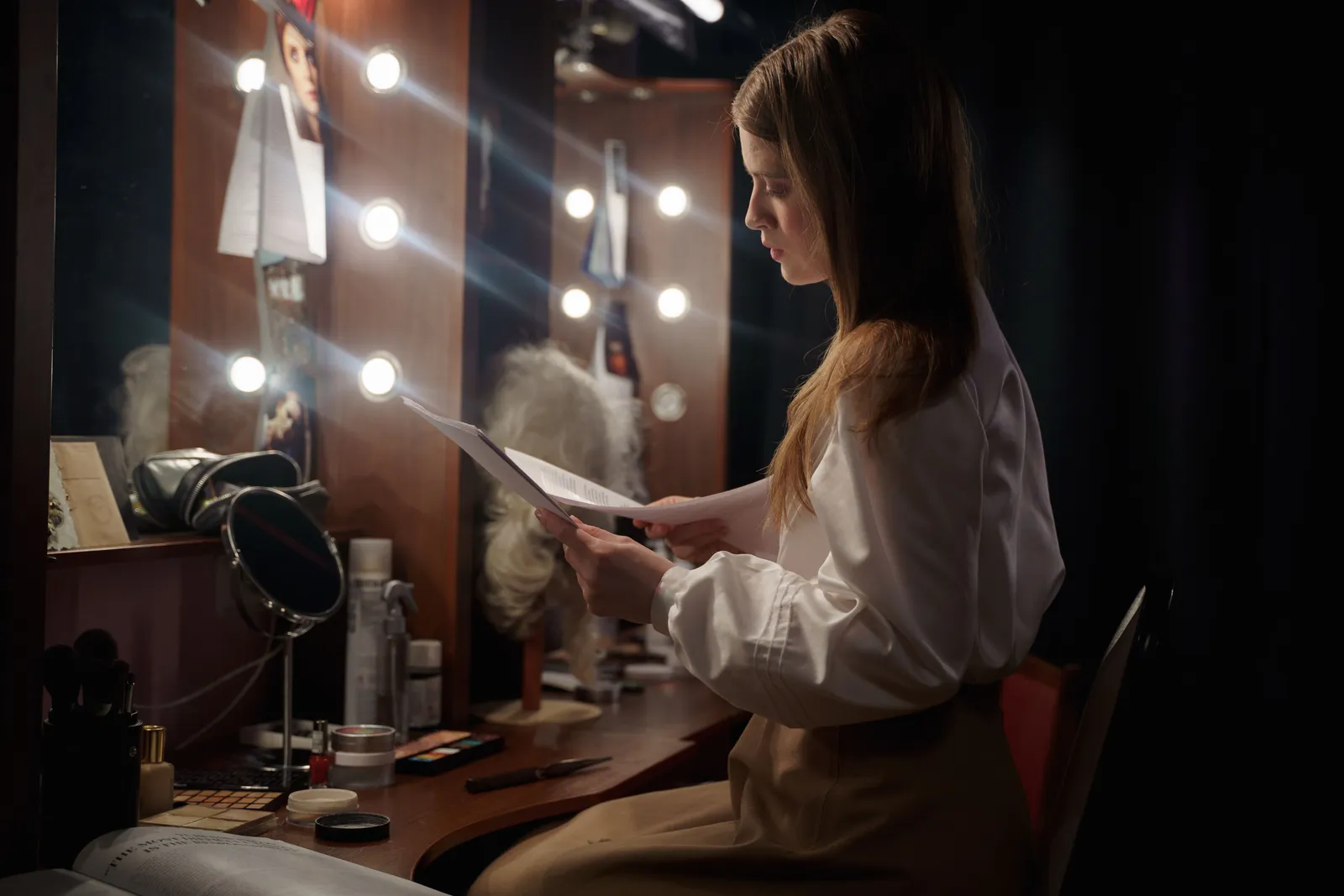 A woman in a dressing room reading a script, surrounded by makeup and lights