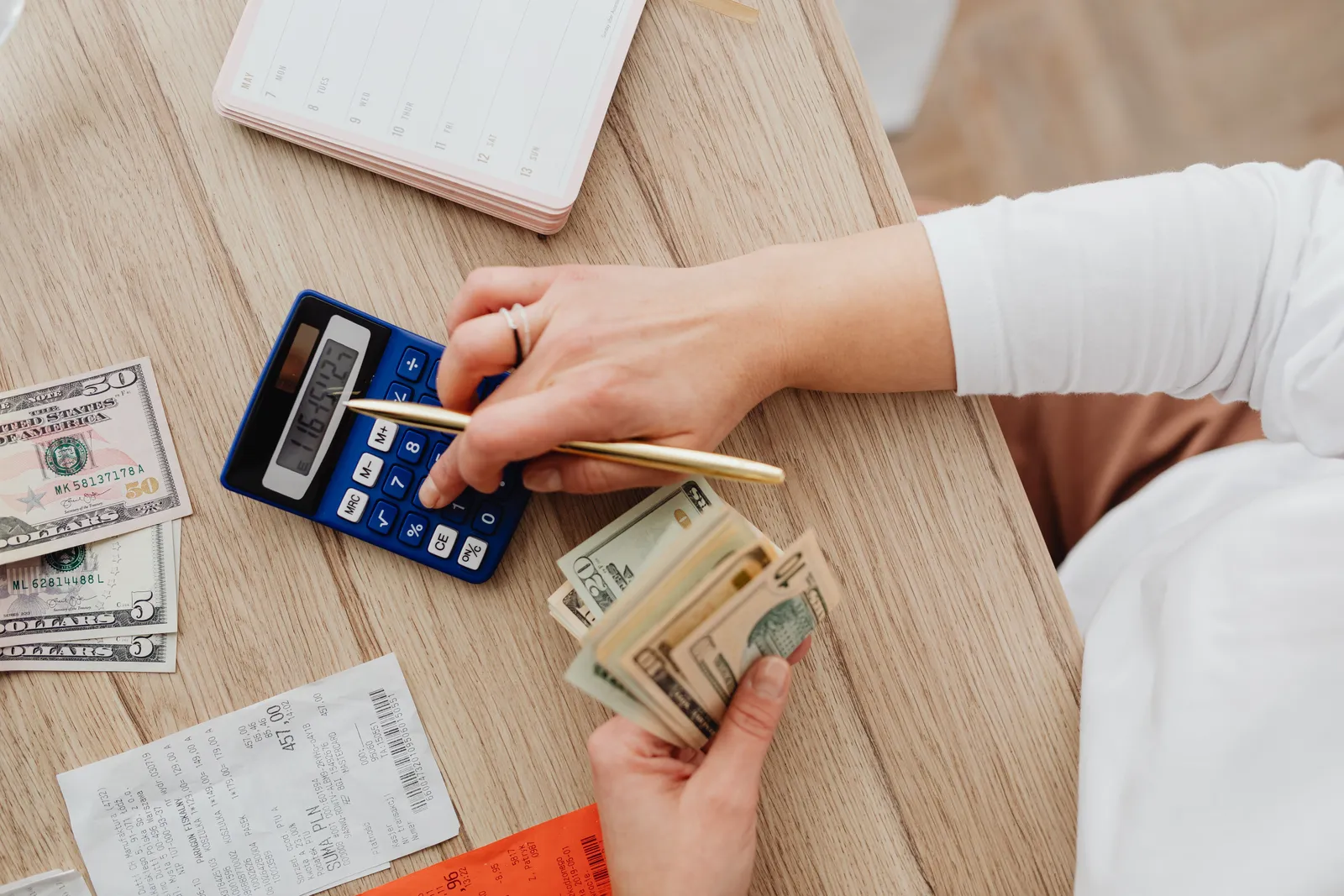 Person using a calculator and counting cash on a wooden table with receipts