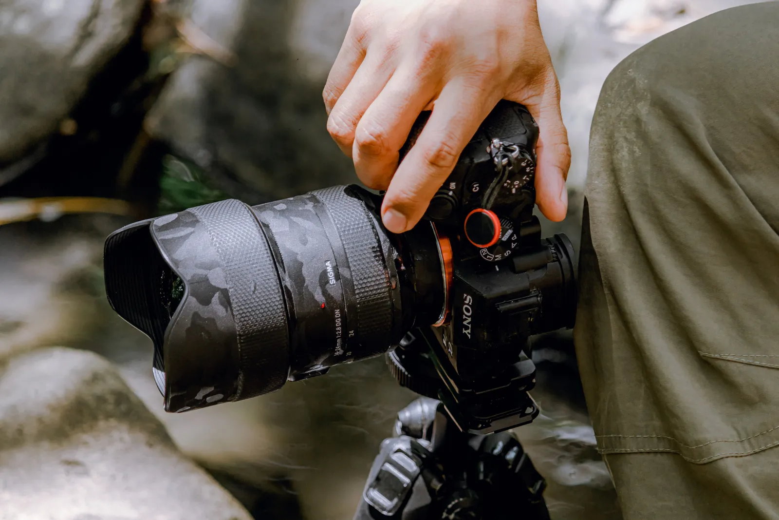 A close-up view of a photographer's hand holding a camera with a camouflage lens cover outdoors