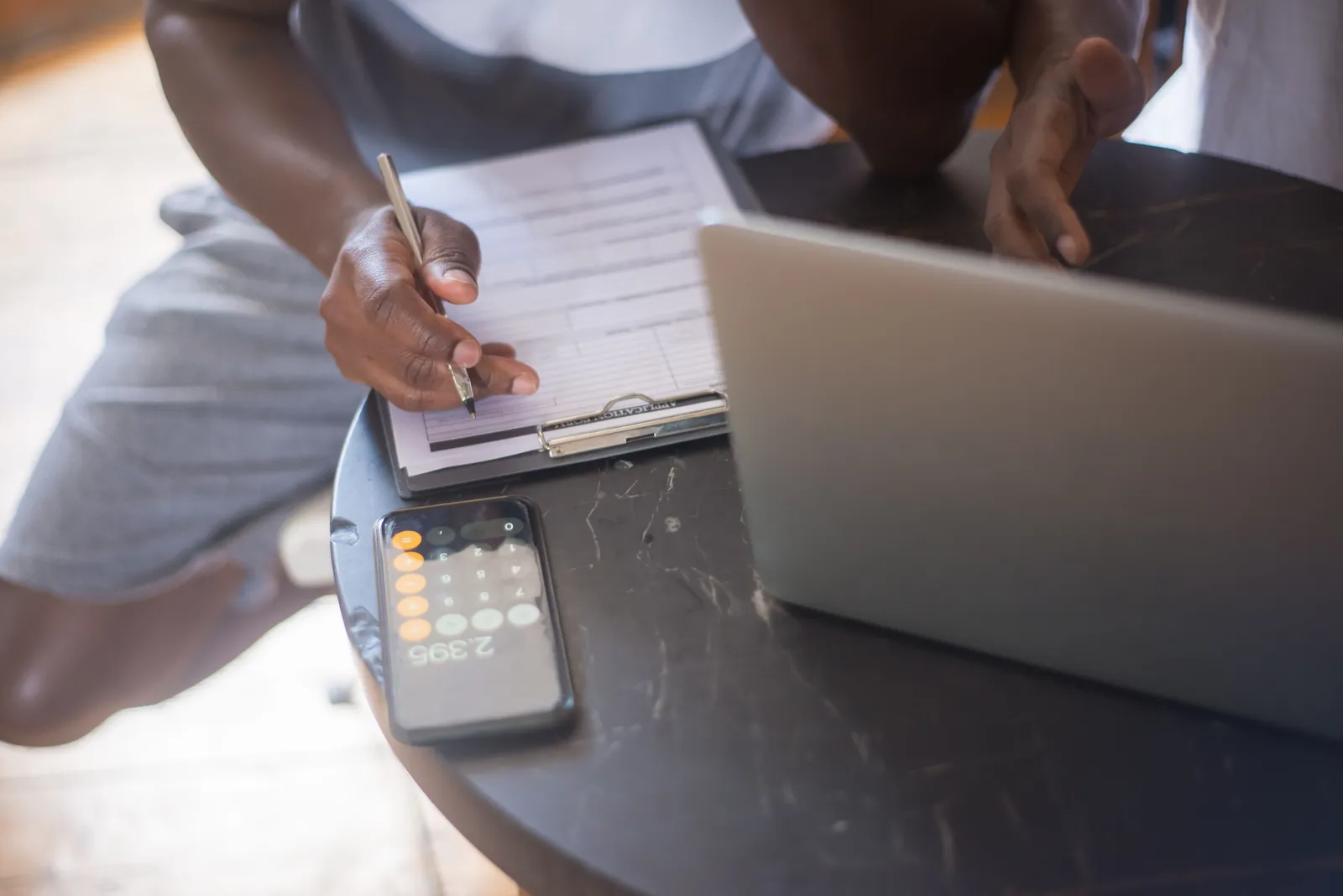 A person working on a laptop with a smartphone and clipboard on a round table