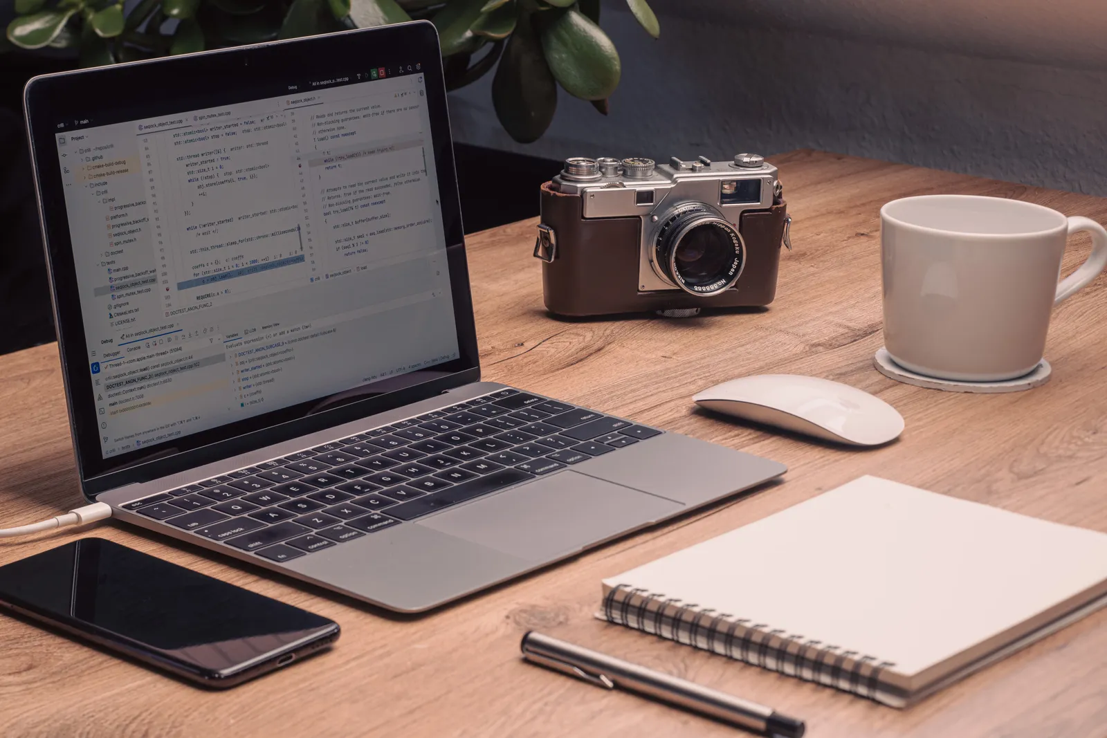 A warm, artistic workspace setup featuring a laptop with visible code, vintage camera, and a notebook on a wooden desk
