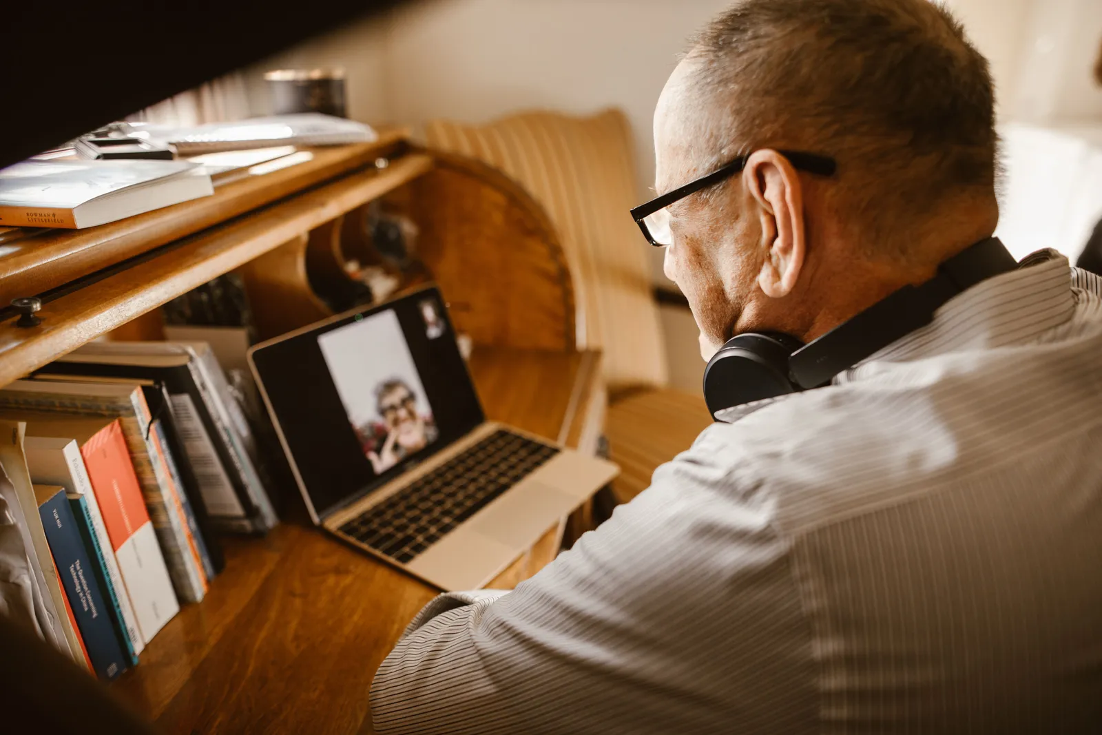 Senior man wearing headphones and eyeglasses on a video call using a laptop at a wooden desk