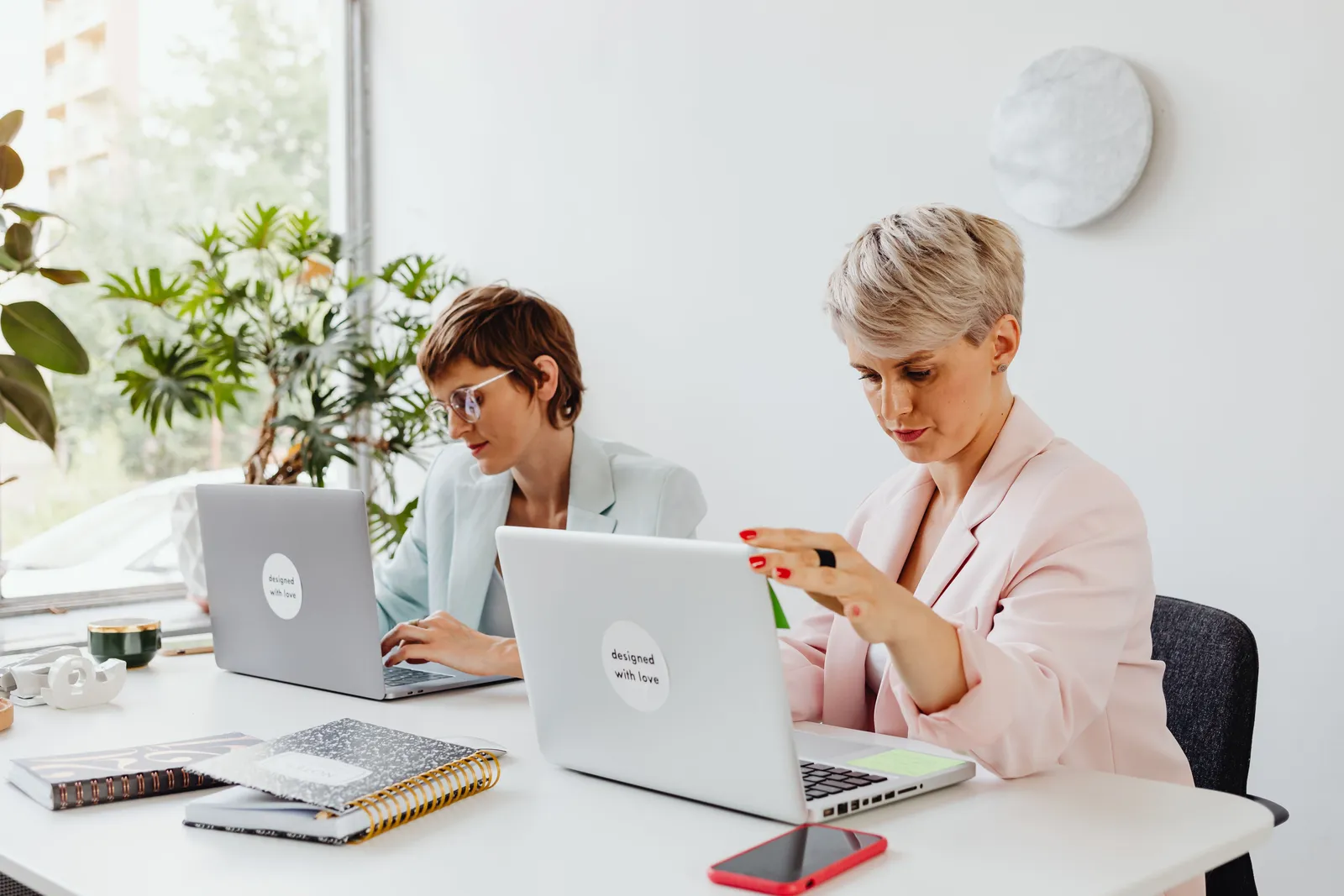 Two businesswomen working collaboratively on laptops in a bright, modern office setting with natural light