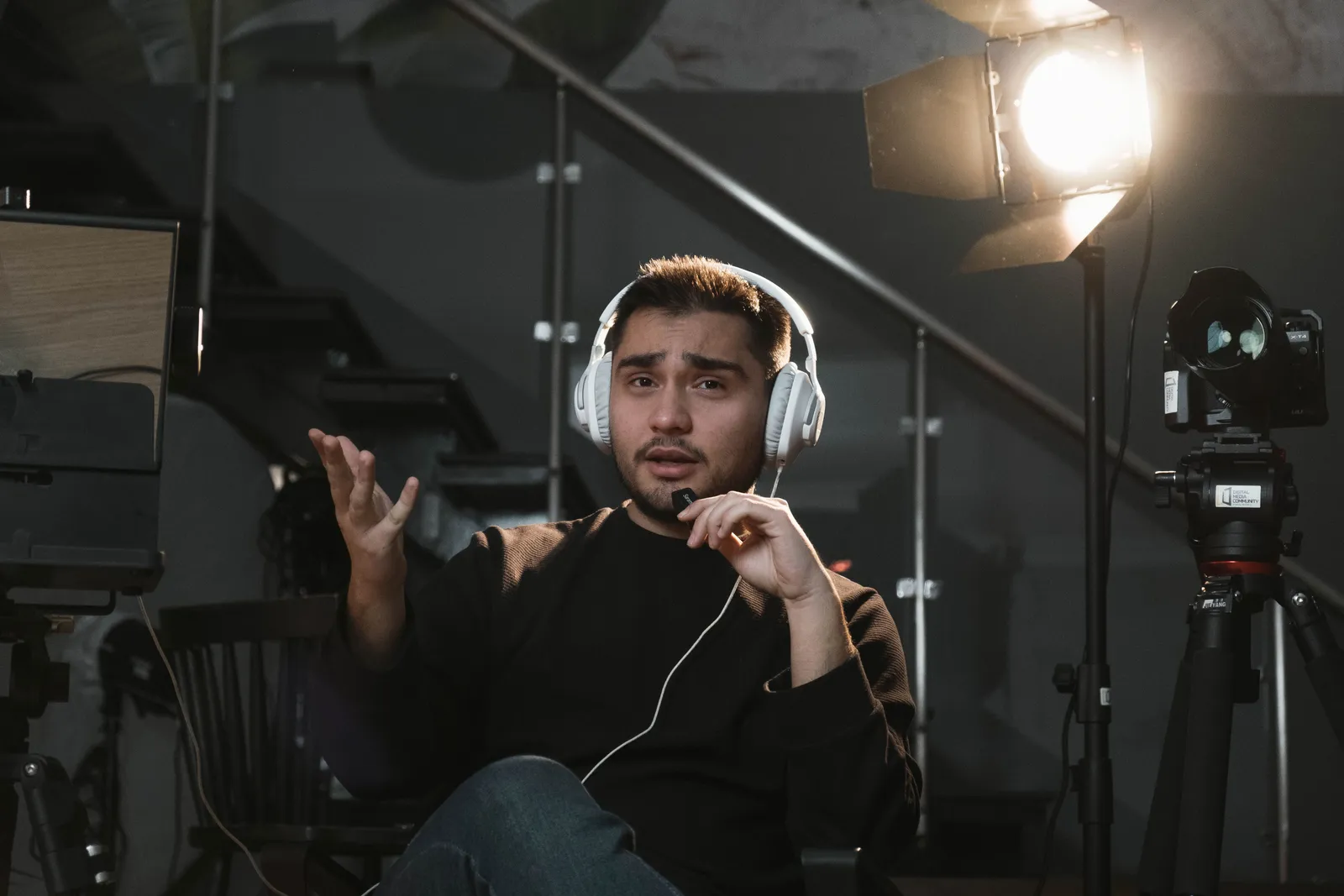 Portrait of a man wearing headphones, sitting in a studio, gesturing towards the camera, with a microphone and lighting setup