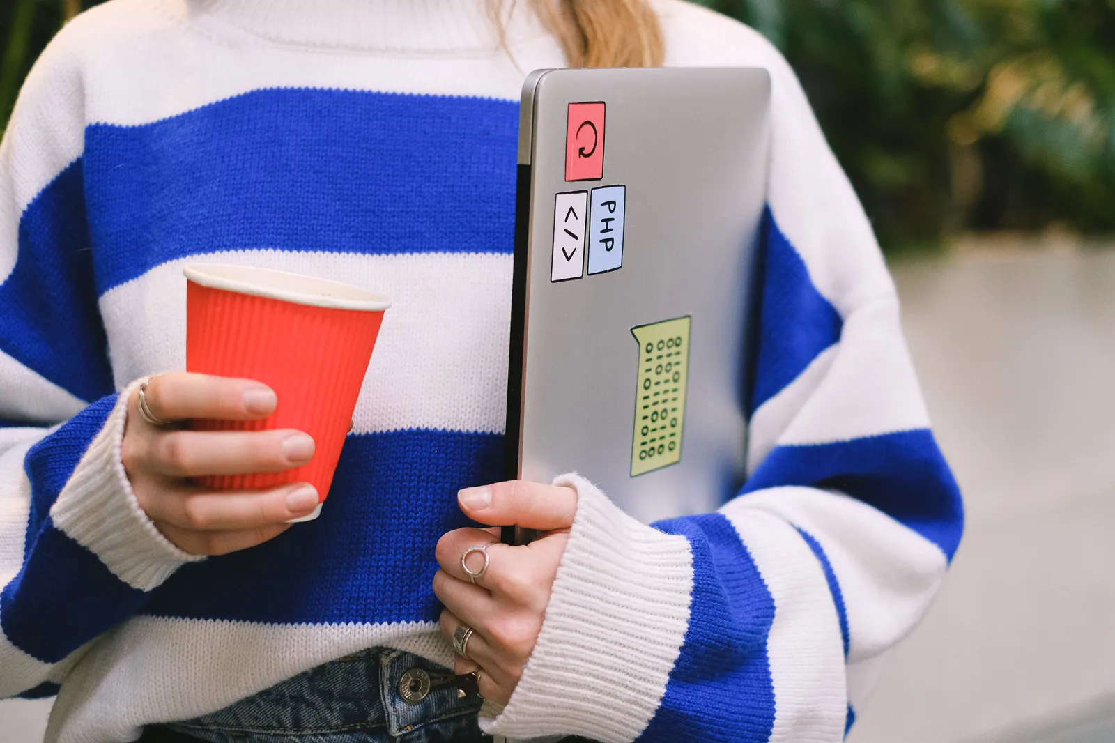Person wearing striped sweater holding laptop with stickers and a red coffee cup indoors
