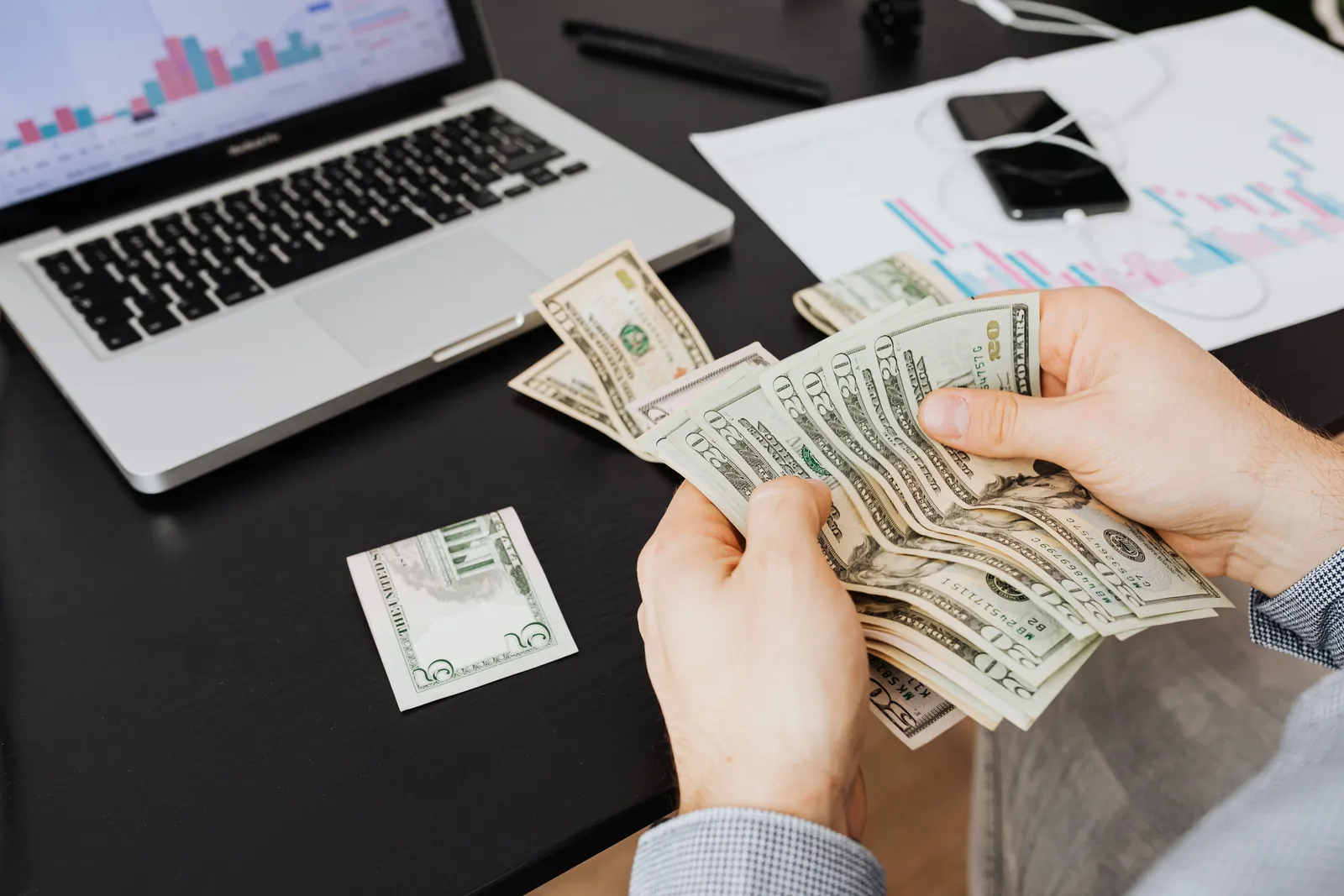 Person counting dollar bills at desk with laptop and financial charts, symbolizing finance management