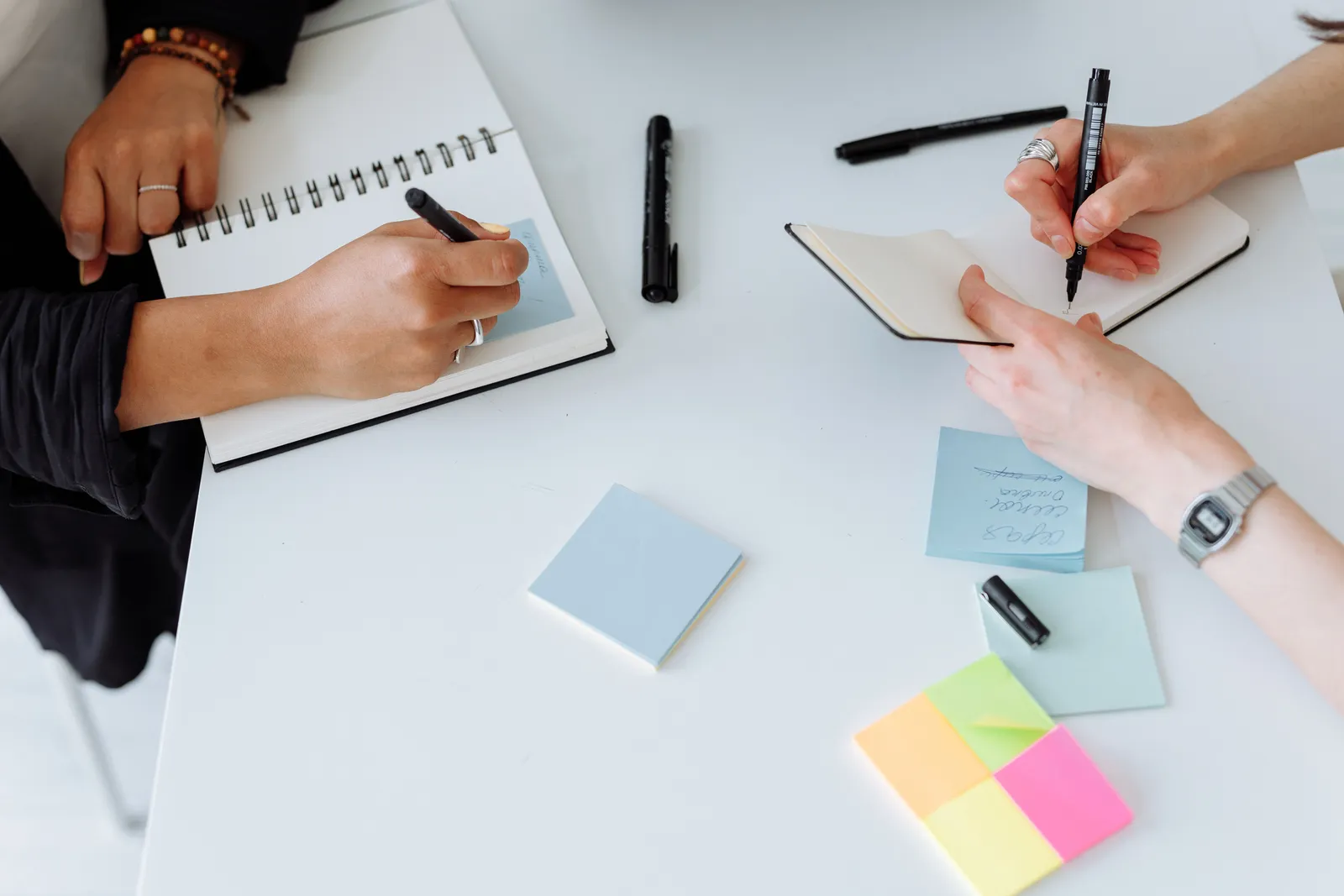 Top view of people collaboratively writing notes on a desk with notebooks and sticky notes