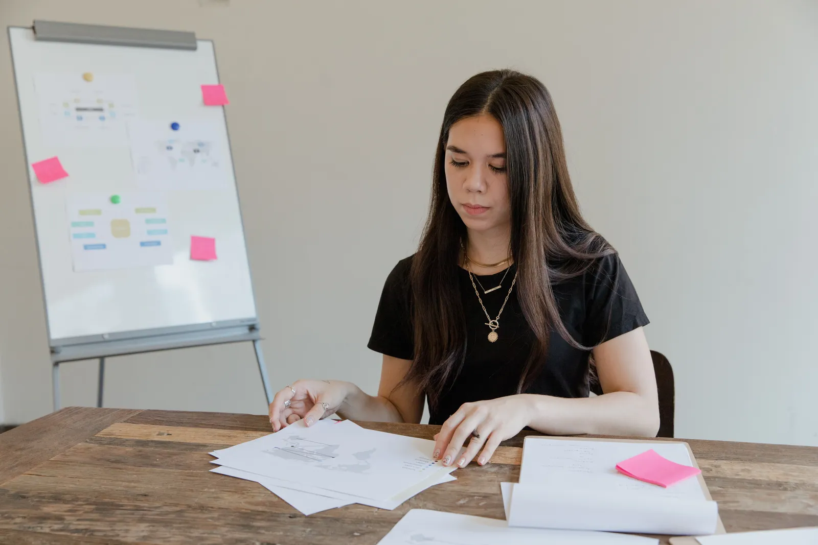 A young woman in a black shirt organizing documents on a wooden table in an office with a whiteboard