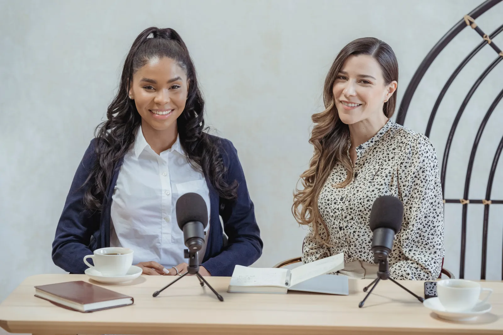 Cheerful multiracial females wearing formal clothes sitting at desk with microphones and looking at camera during interview